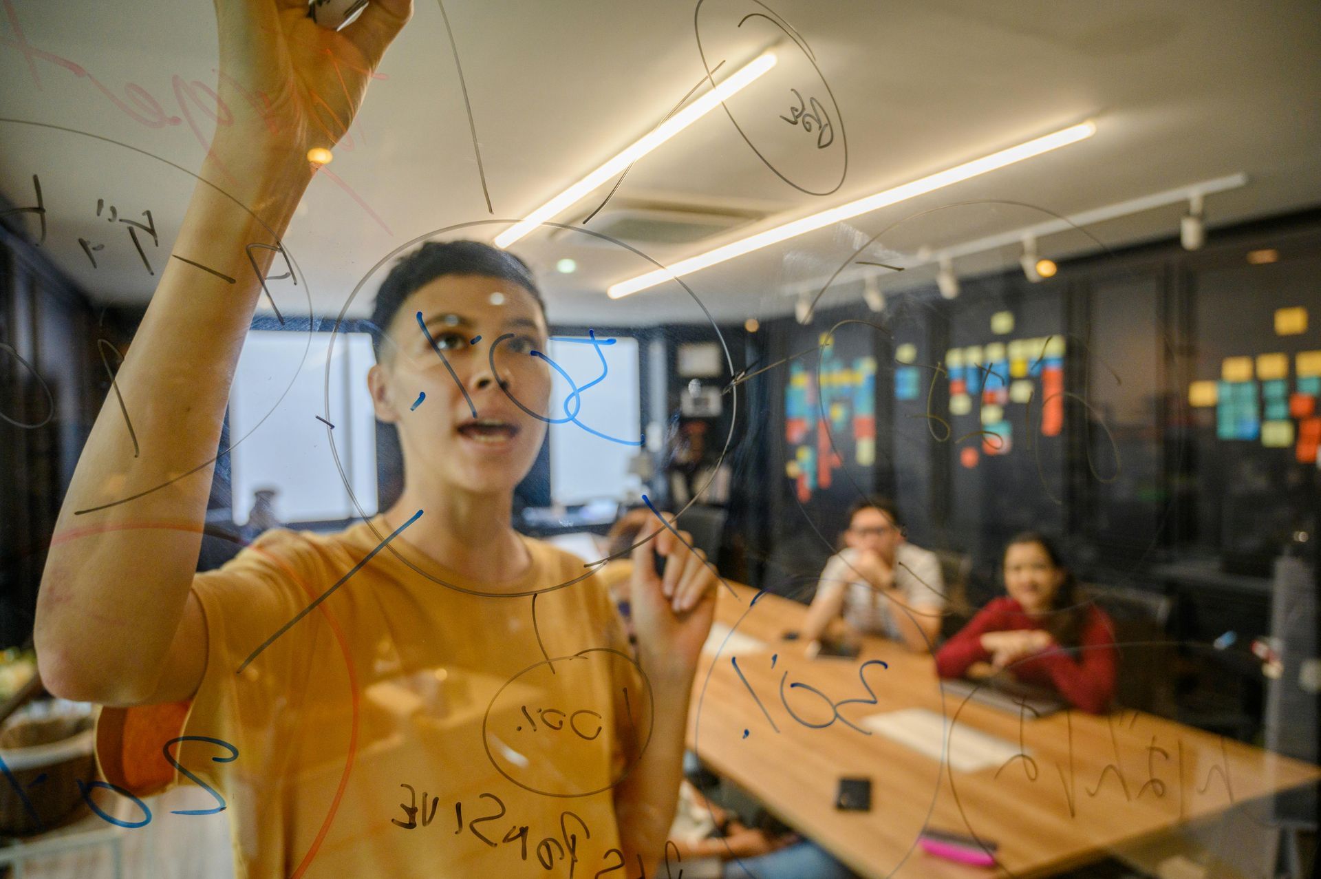 A person writes on a glass board in an office while two colleagues sit at a table in the background.