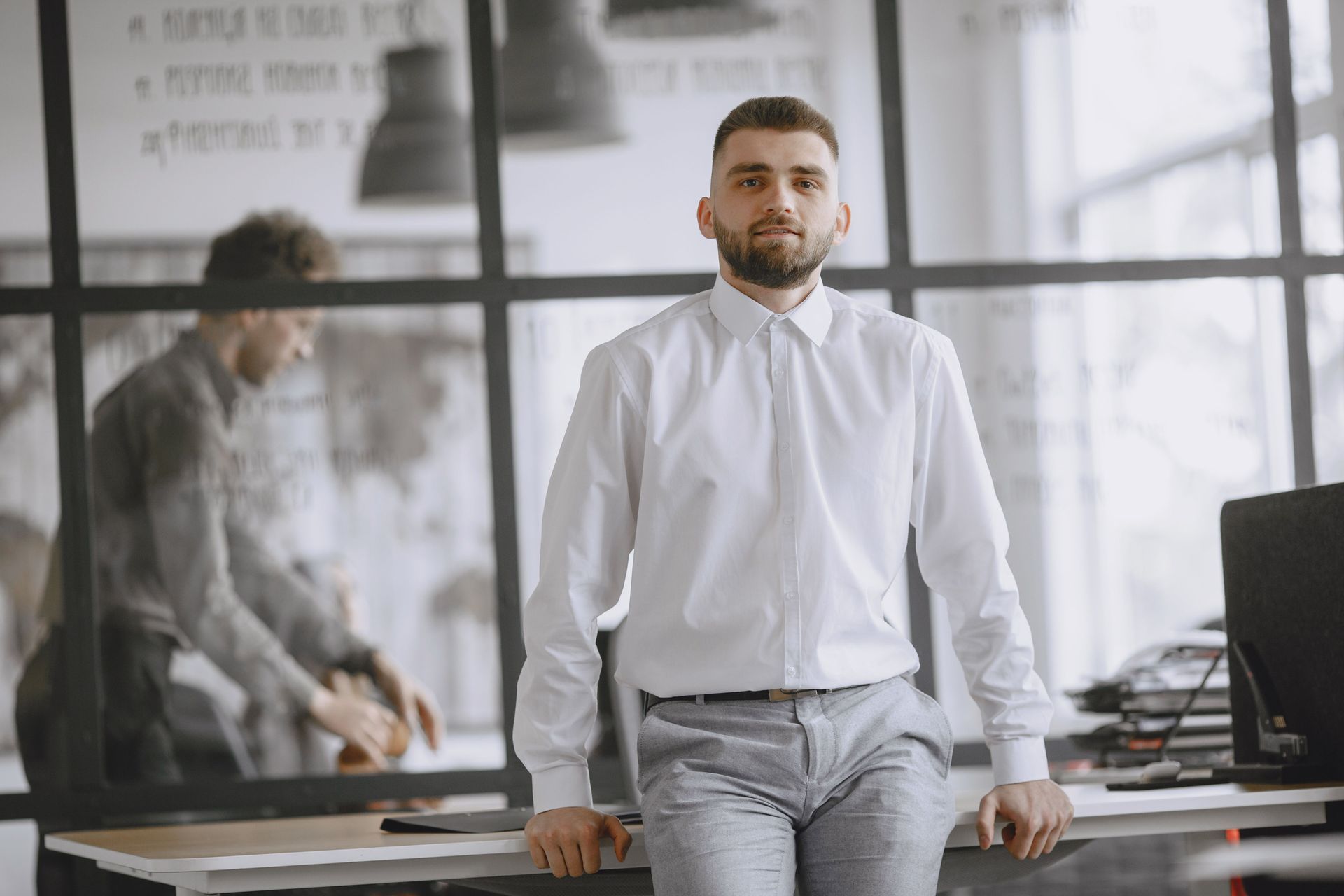 Man in a white shirt leaning on a desk in a modern office, with a coworker blurred in the background.