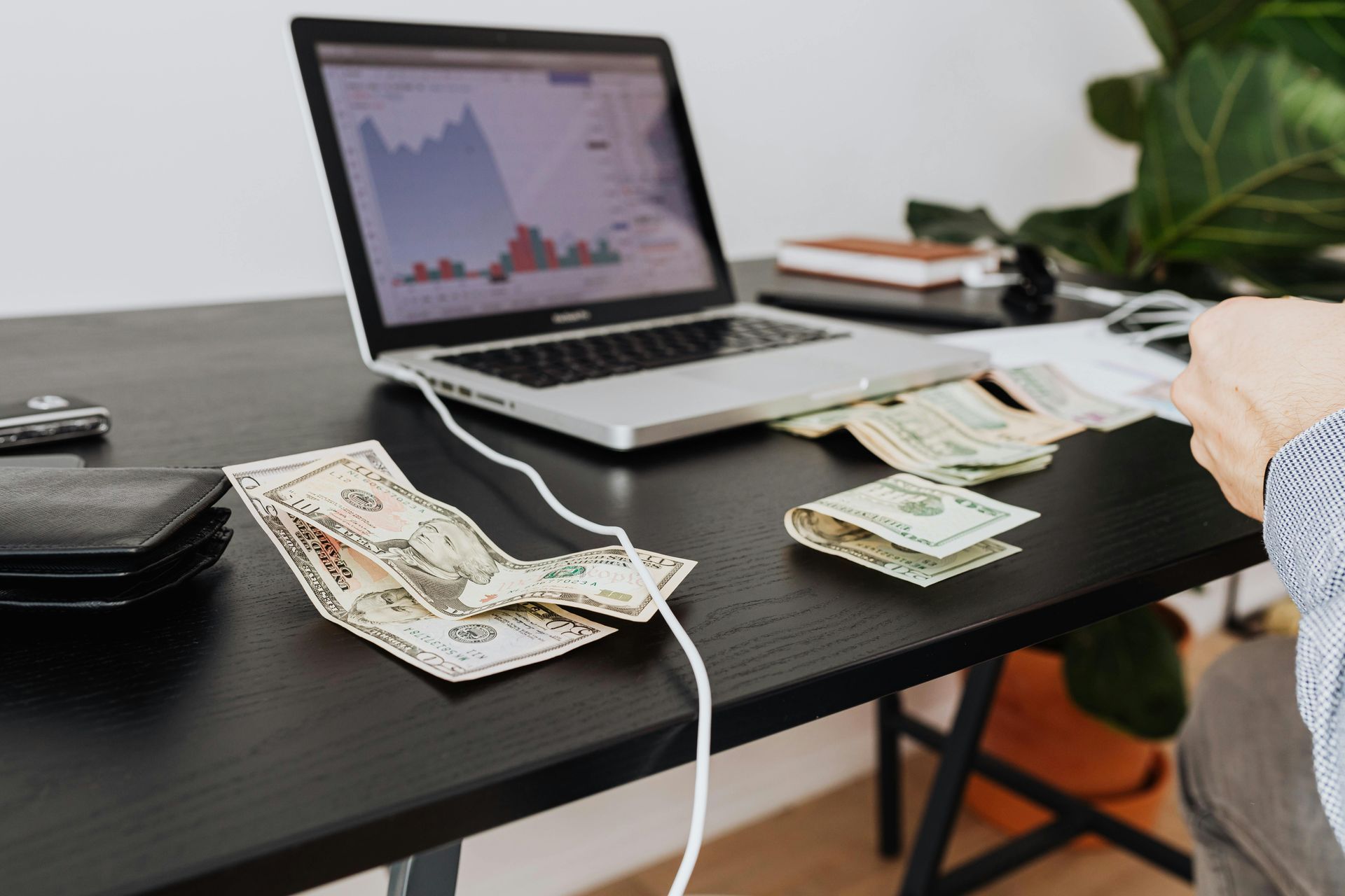 Laptop on dark desk with currency, charging cable, and a hand counting money by papers and books