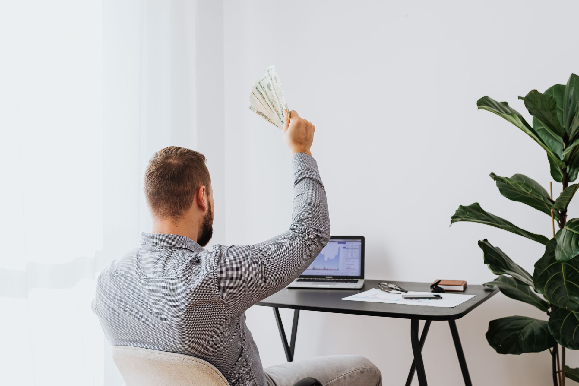Person at a desk holding up cash near a laptop and monitor, in a bright home office.