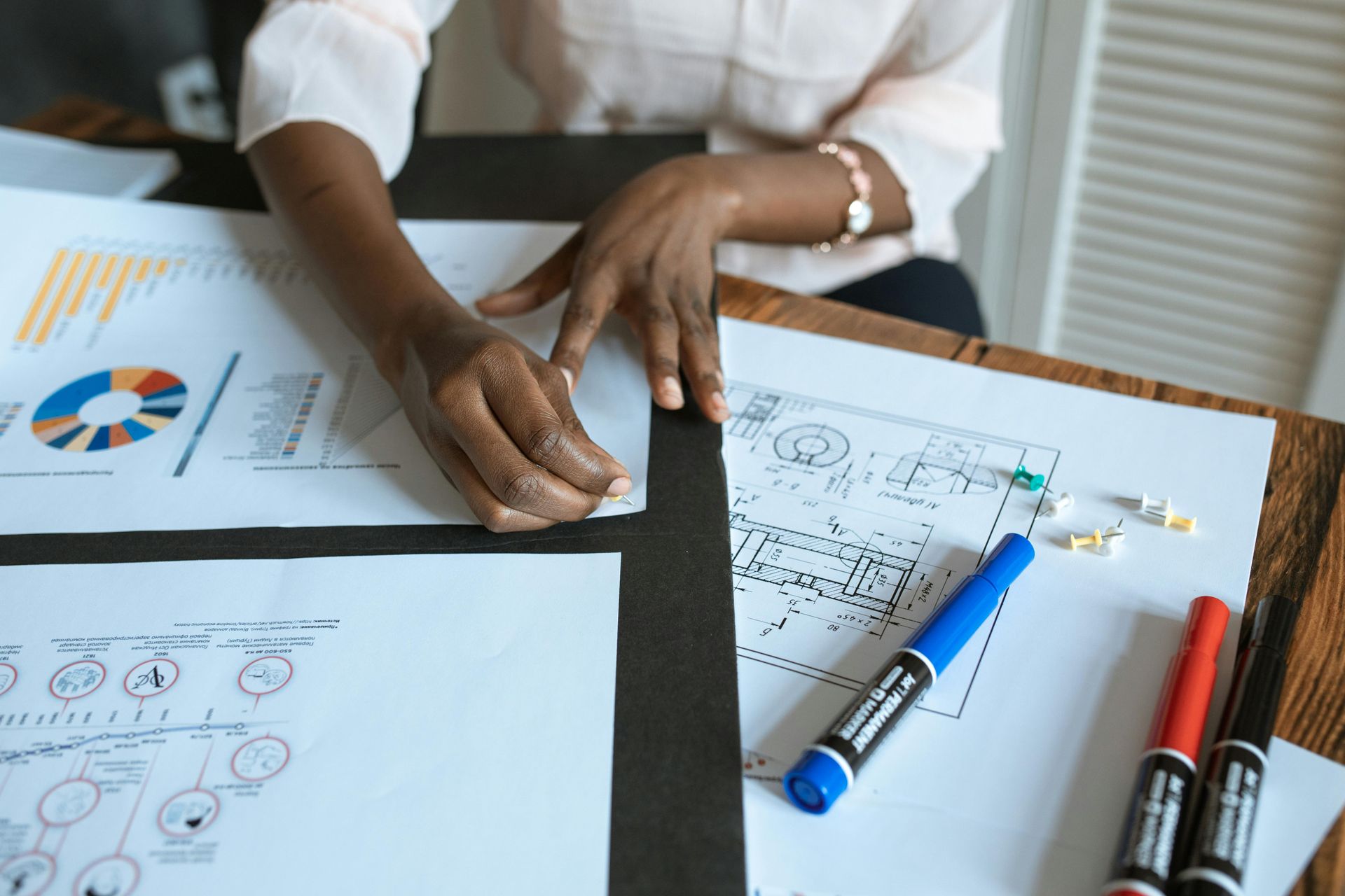 A person sits at a wooden table reviewing charts and technical floor plans with markers nearby.