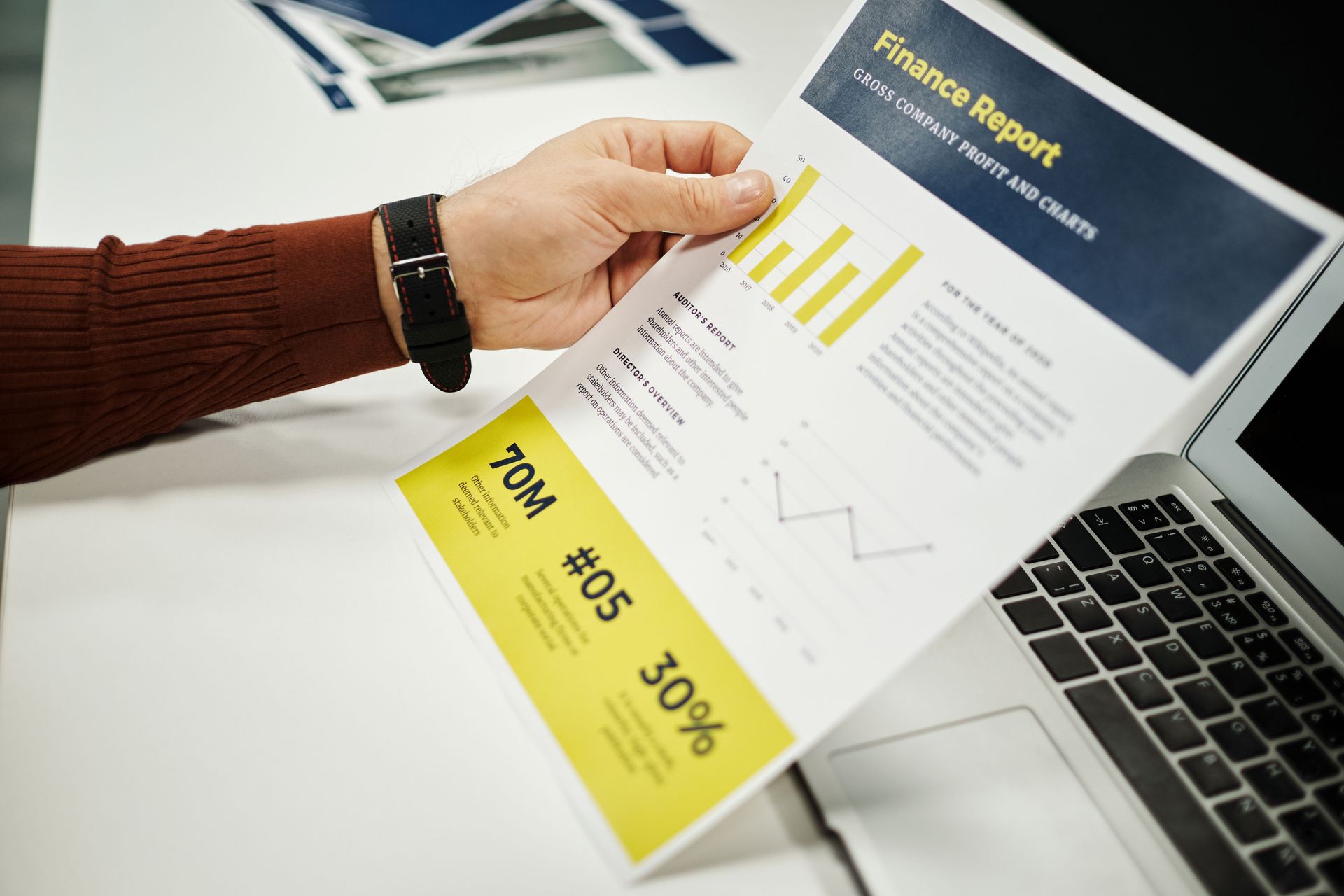 Hand holding a financial report with charts and a yellow summary sheet over a laptop keyboard