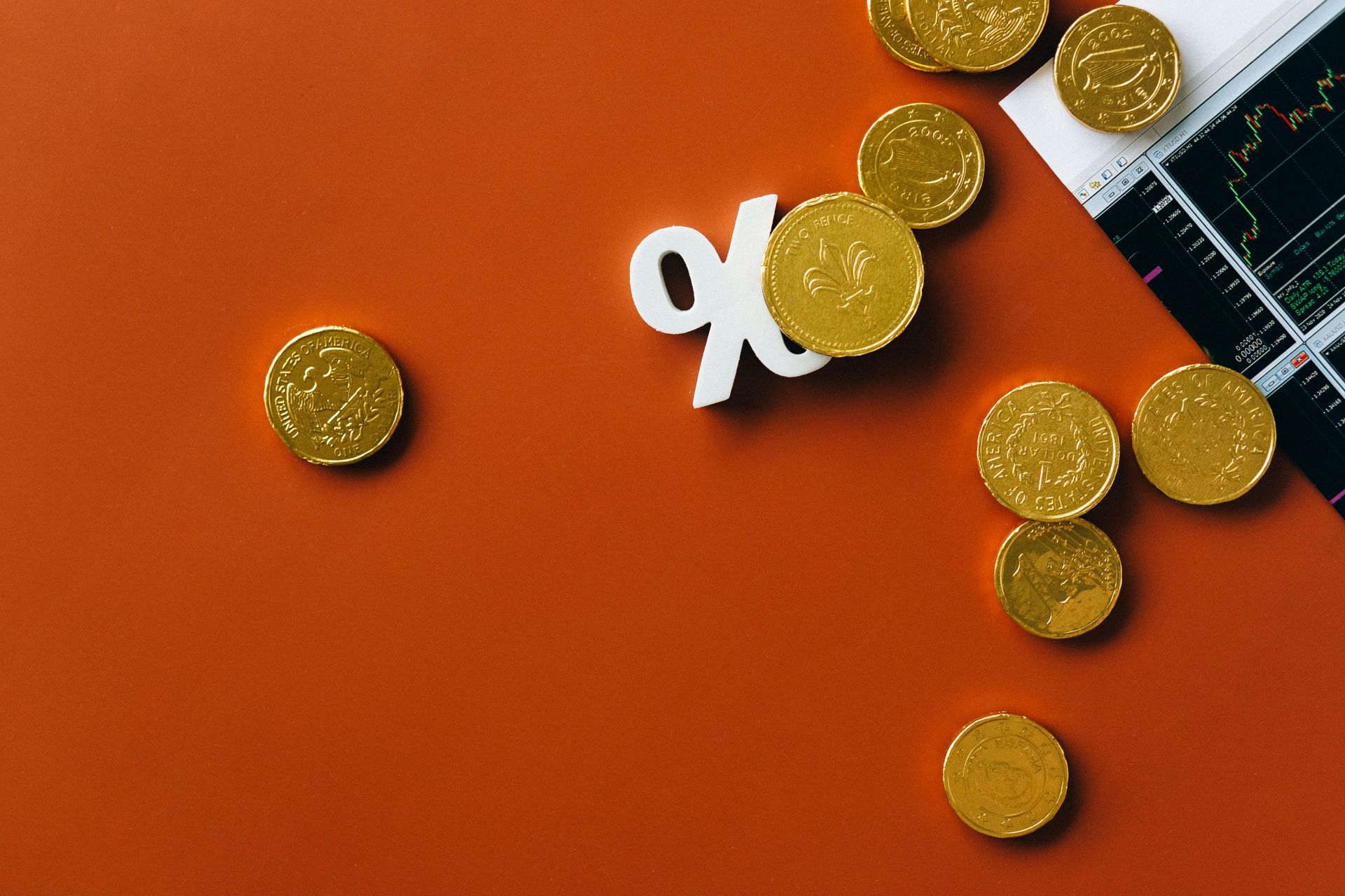 Gold coins and a percentage symbol arranged on an orange background next to a financial stock chart.
