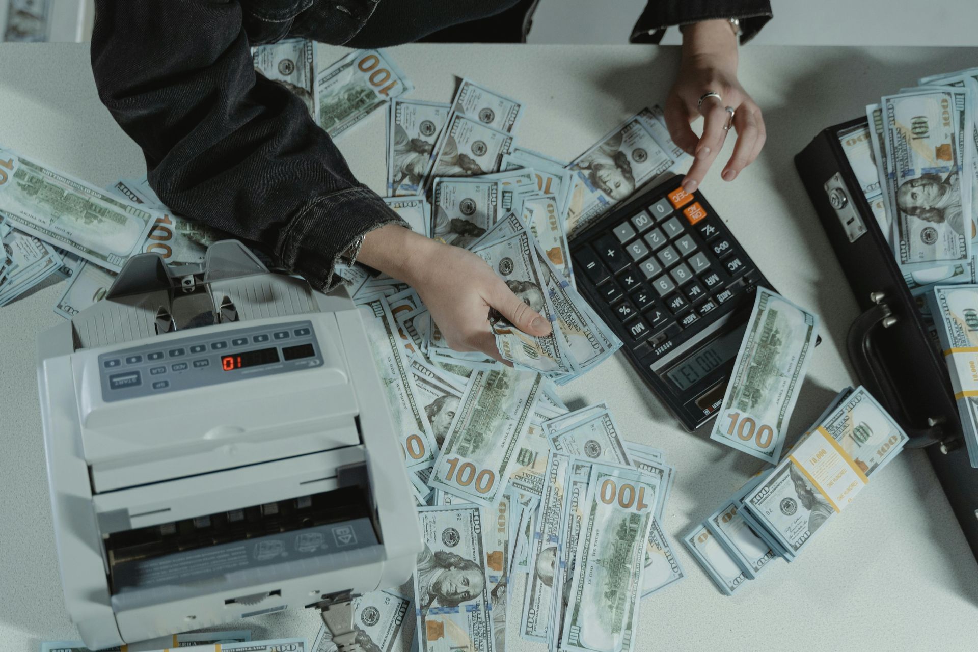 A person uses a calculator and a money-counting machine on a desk covered with numerous stacks and loose $100 bills.