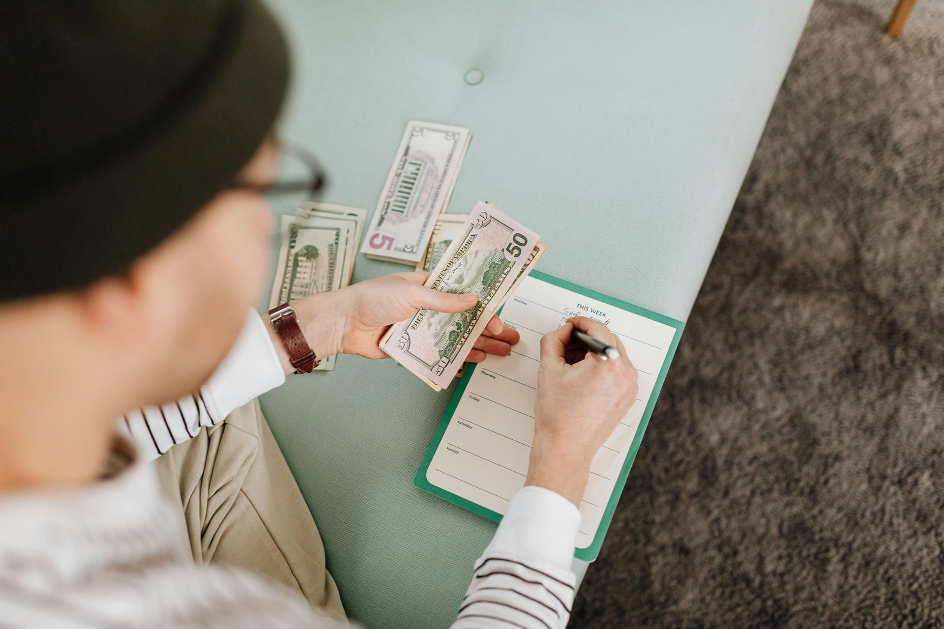 A person sitting on a blue sofa holds stacks of US dollar bills while writing in a notebook.