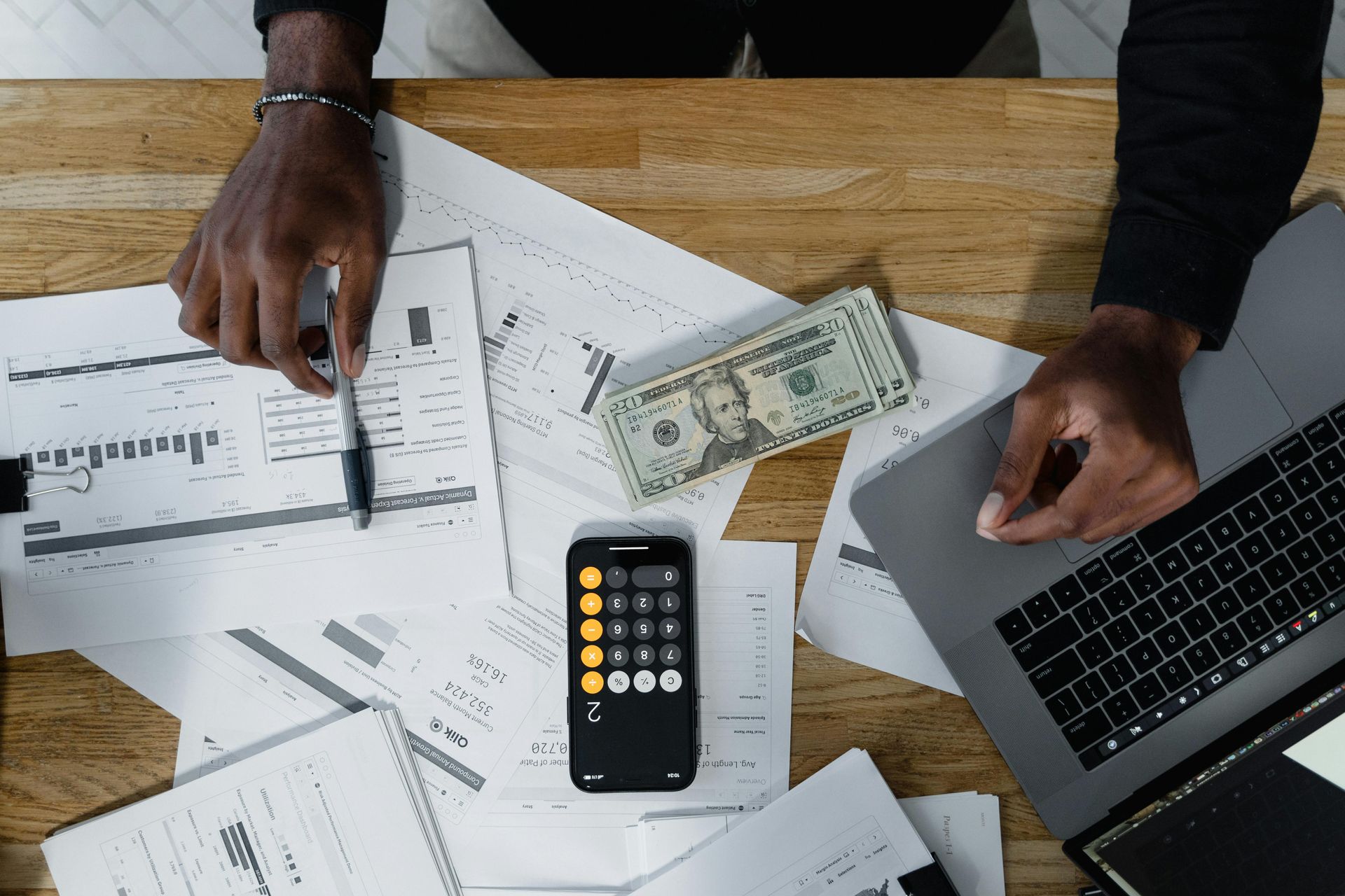A person working at a wooden desk with a laptop, calculator, documents, and a stack of US currency.