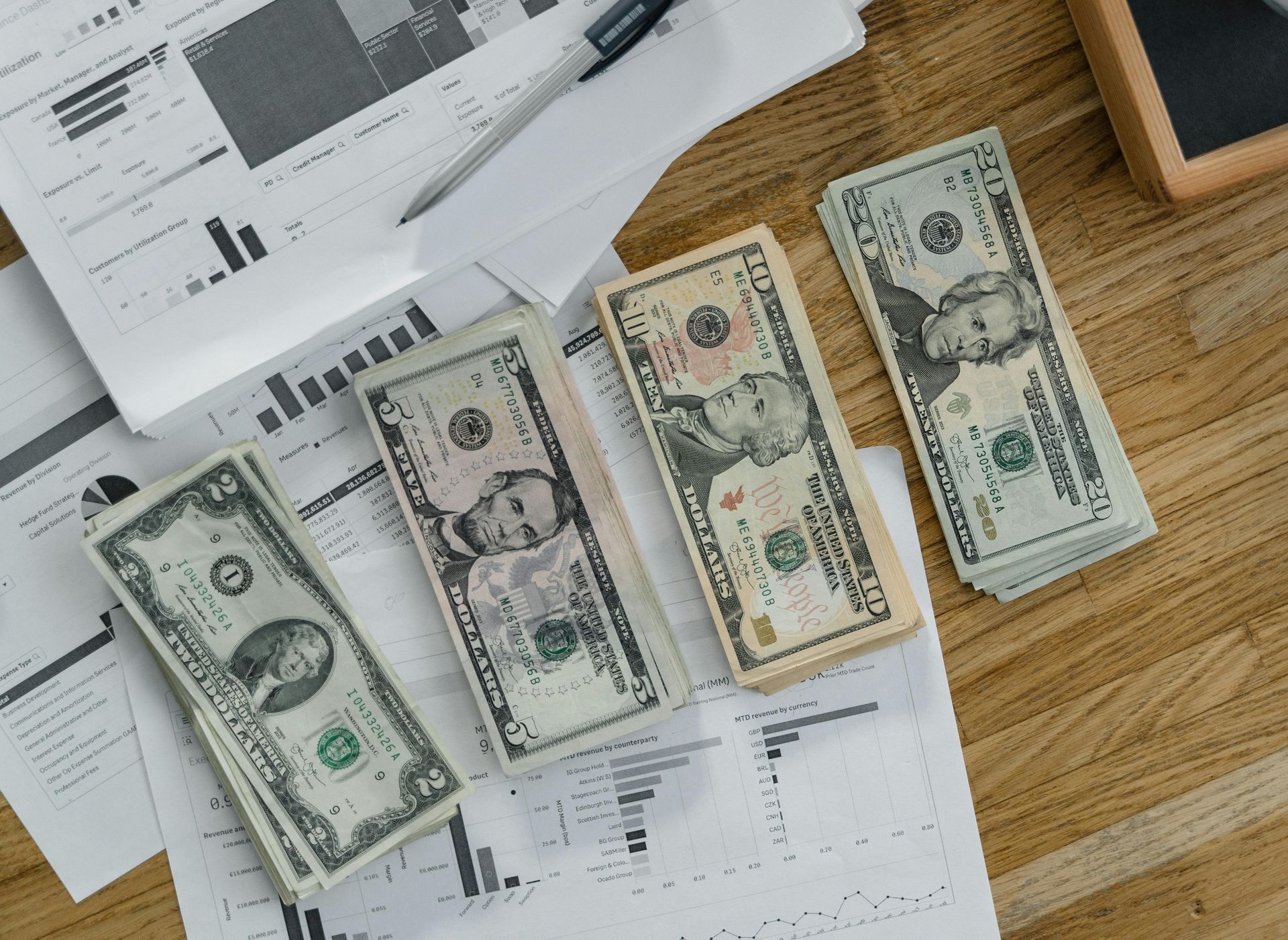 Three stacks of US dollar bills placed on top of financial spreadsheets and a pen on a wooden desk.