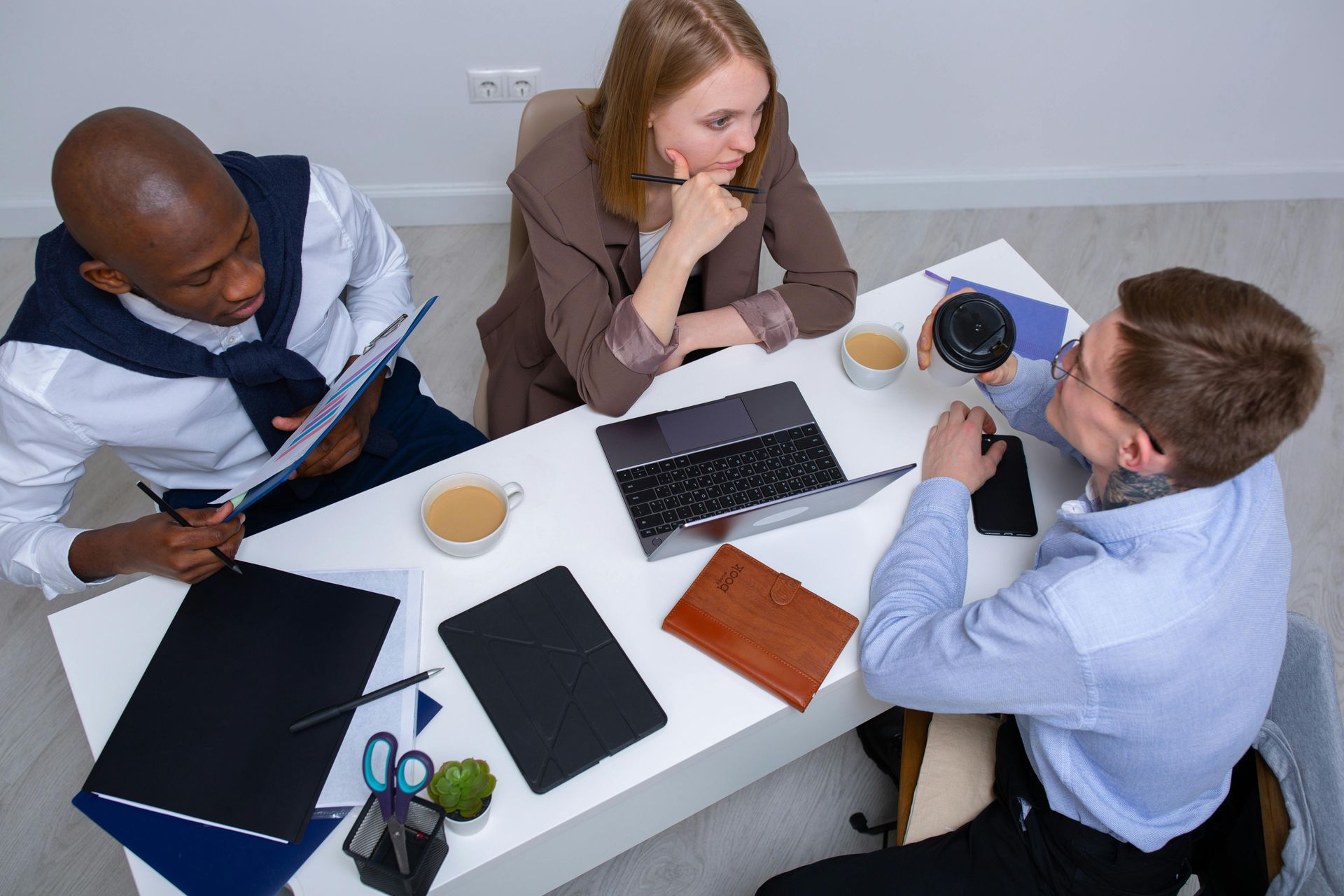 Three professionals sit around a white desk in a bright office, working together with laptops and documents.