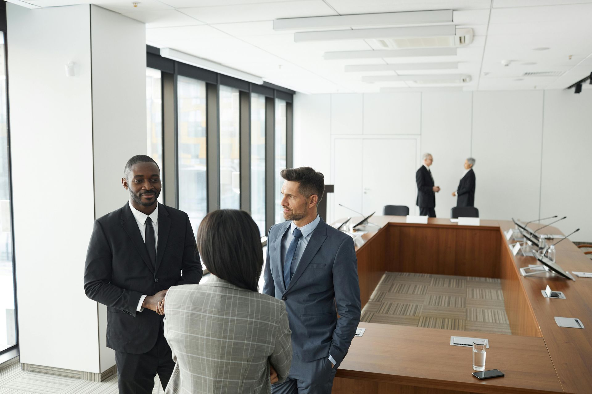 Professional colleagues in suits talking in a bright boardroom with a large wooden table, while two others converse nearby.