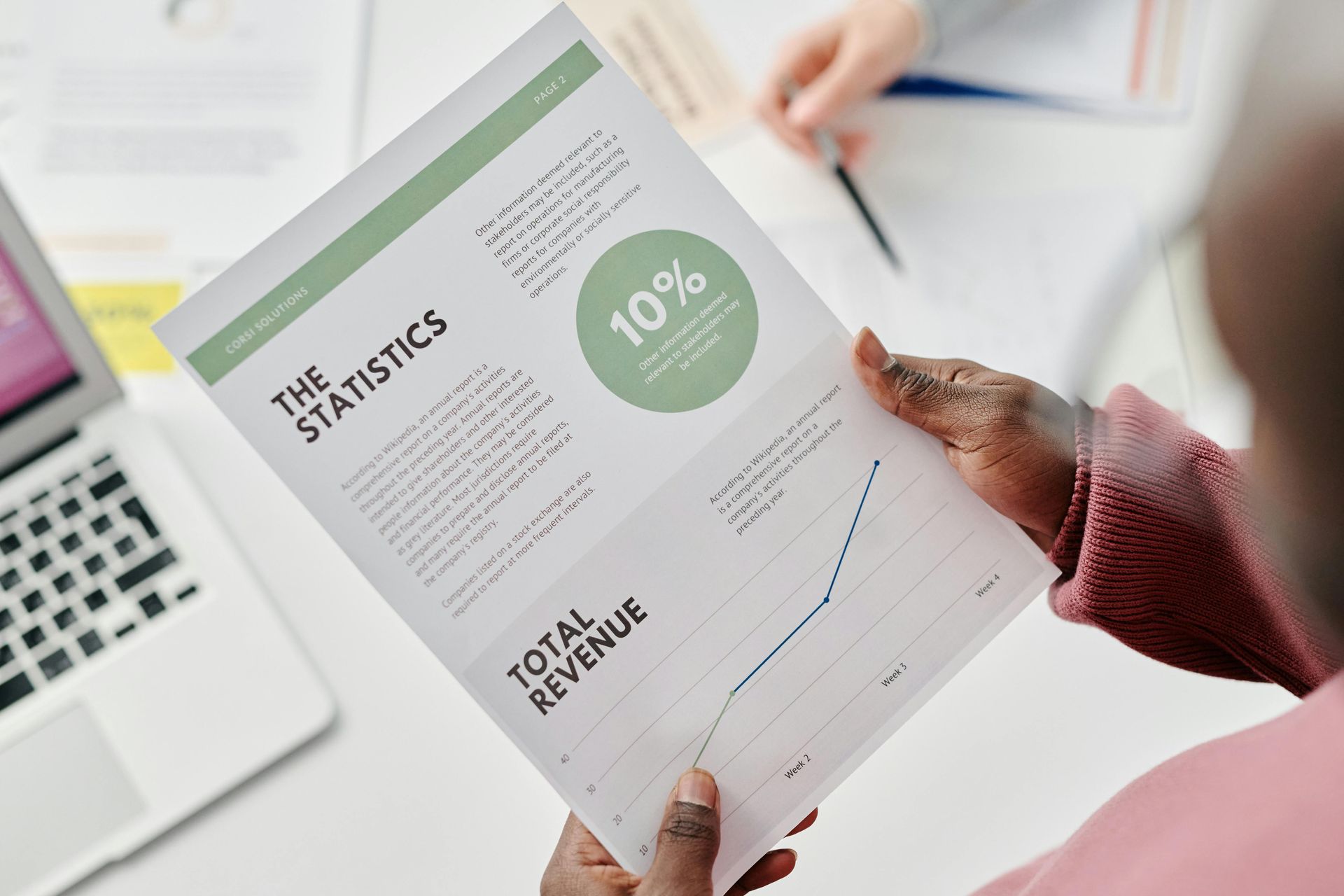 Person holding a document with statistics and a graph, working with another person at a table.