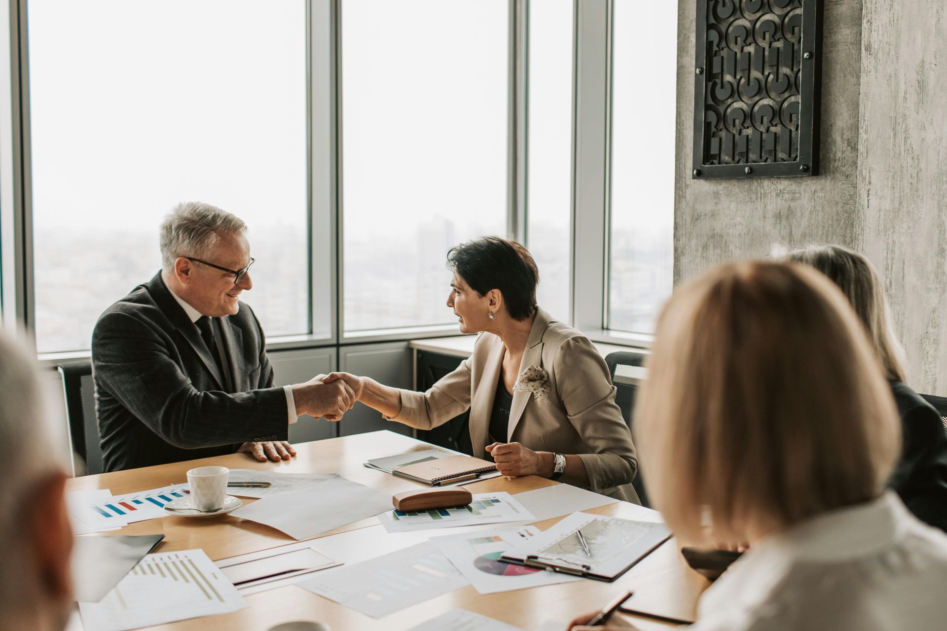 Two professionals shake hands over a meeting table with documents and a city skyline visible through large windows.