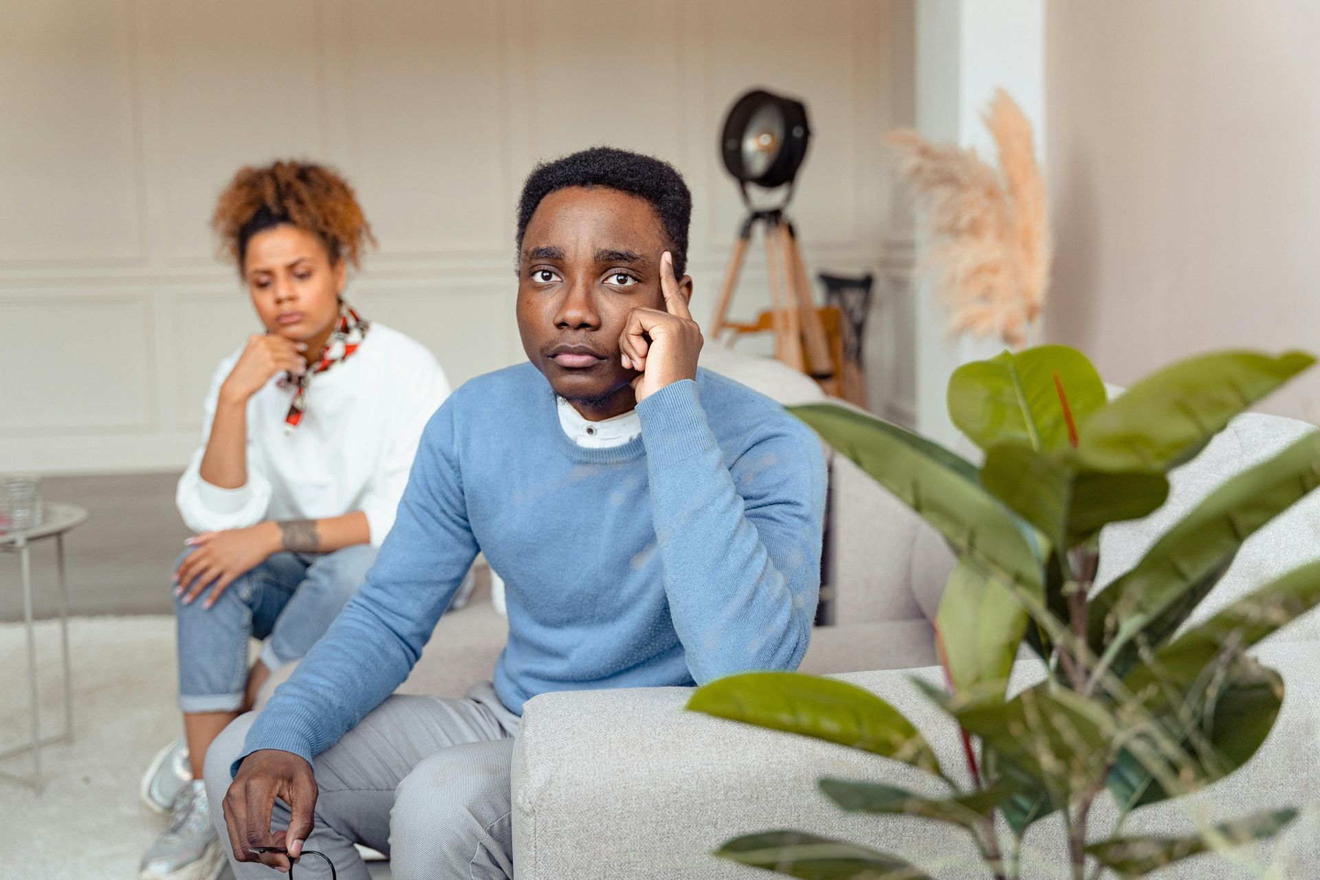 Two people sit on a couch in a bright room, both looking thoughtful and pensive with their hands near their faces.