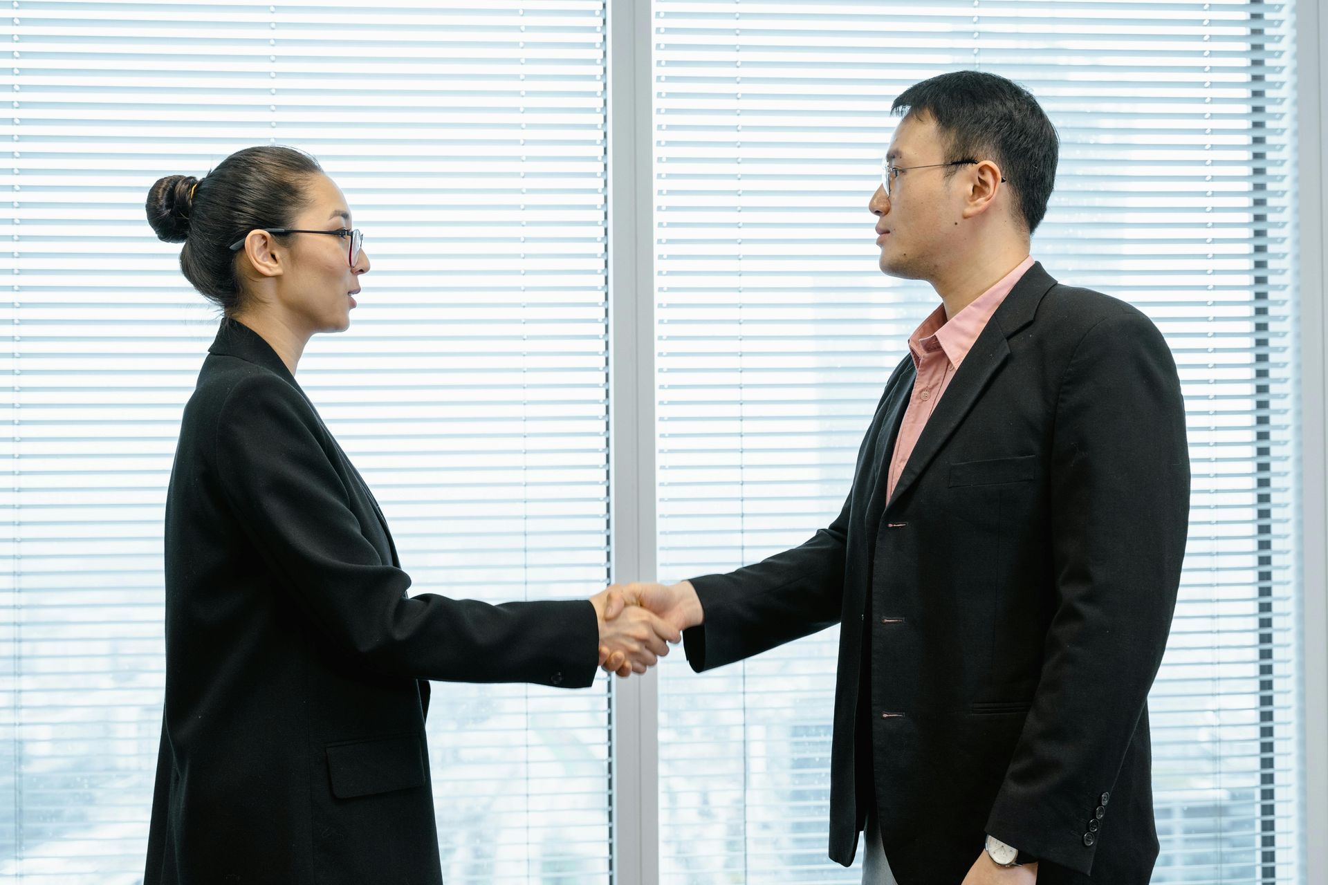 Two professionals in business attire shake hands in front of a window with blinds.