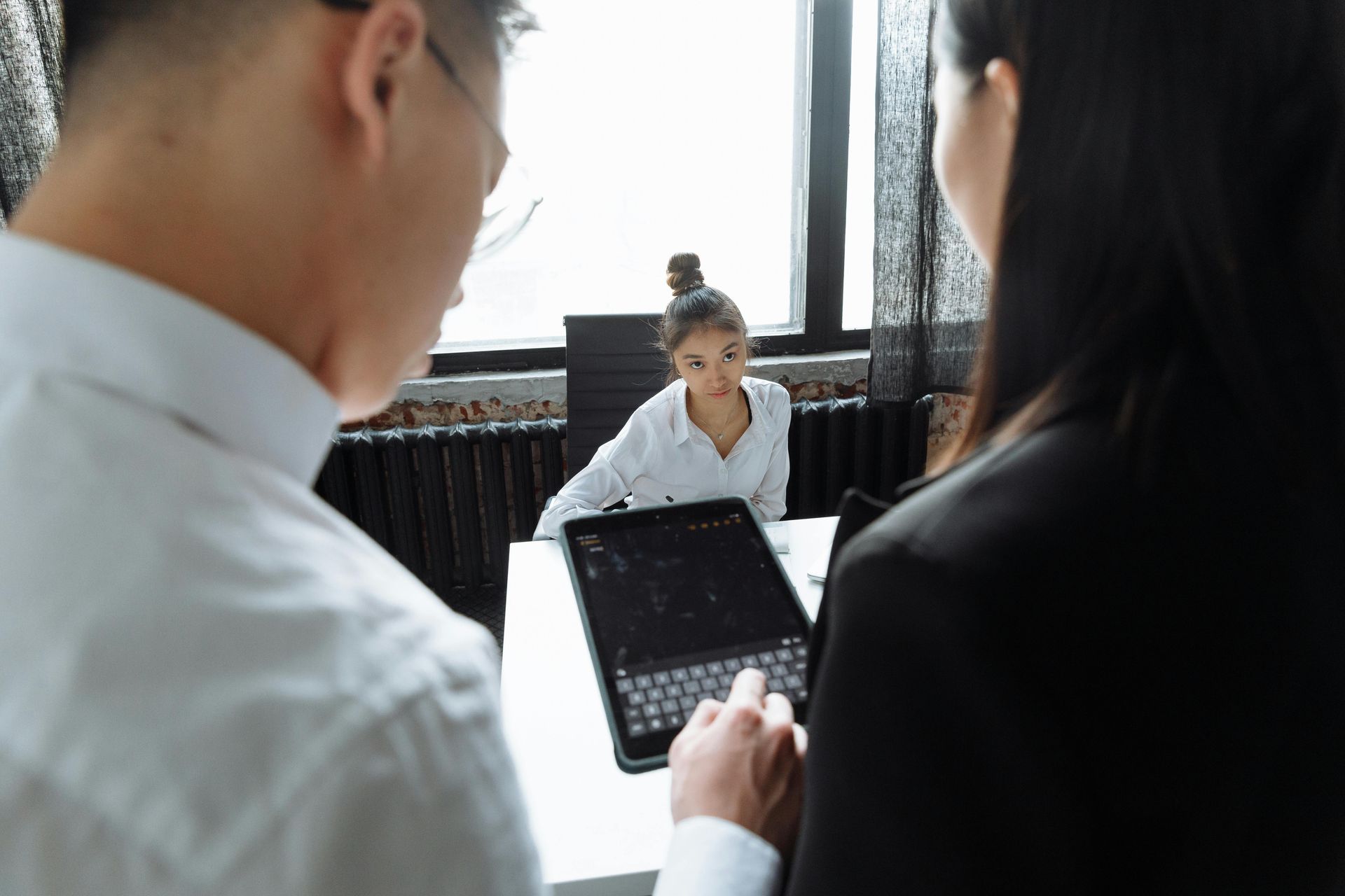 Two people in business attire look at a tablet together while a third person sits at a desk in a bright office.