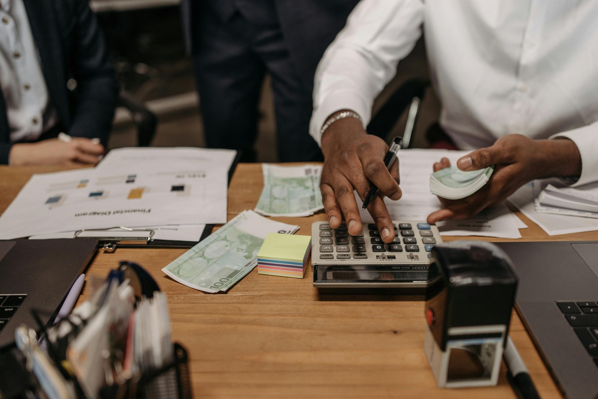 A person uses a calculator on a desk scattered with currency, documents, a stapler, and a laptop in an office setting.