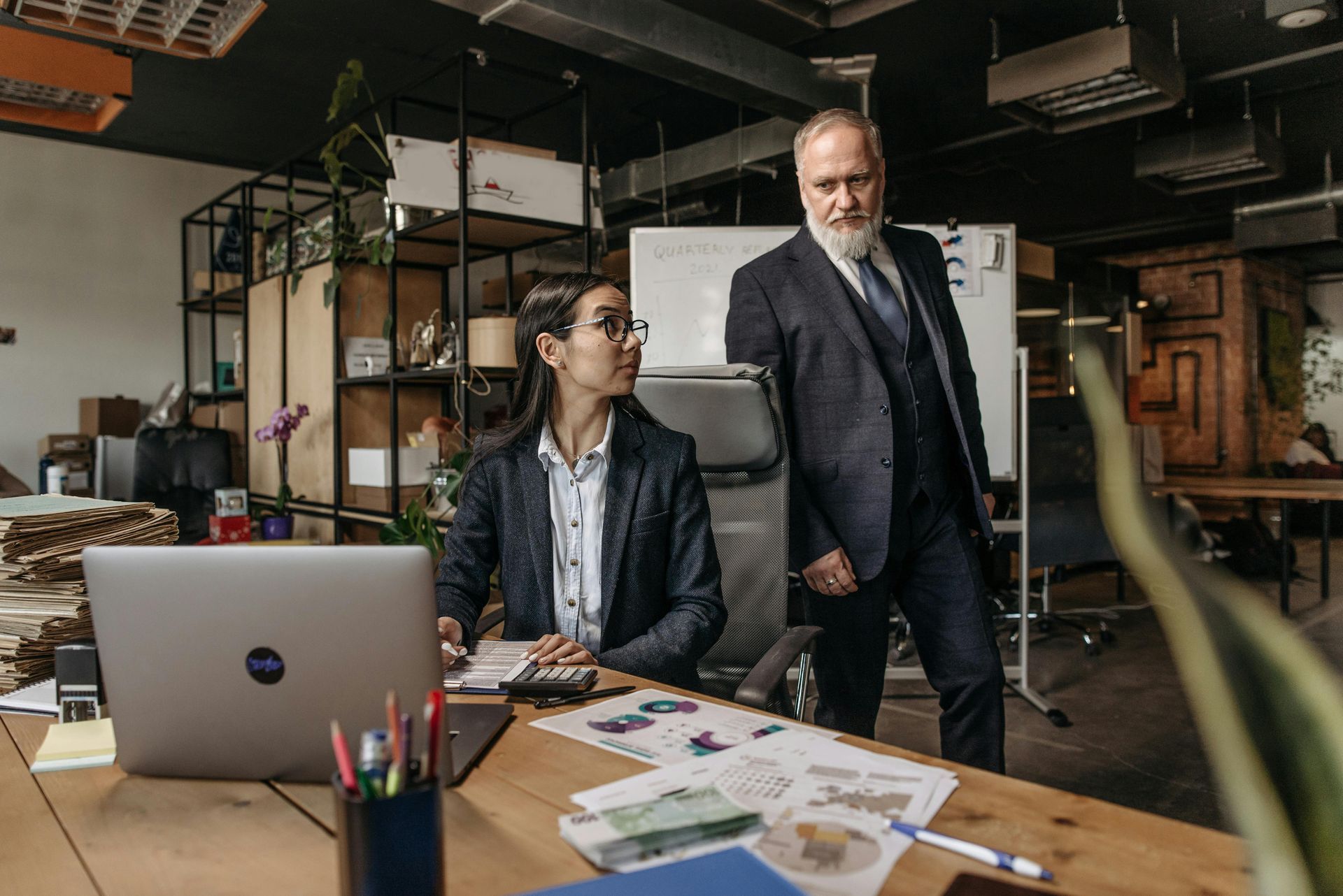 A professional woman works on a laptop at a desk while a man in a suit stands beside her in a modern office.