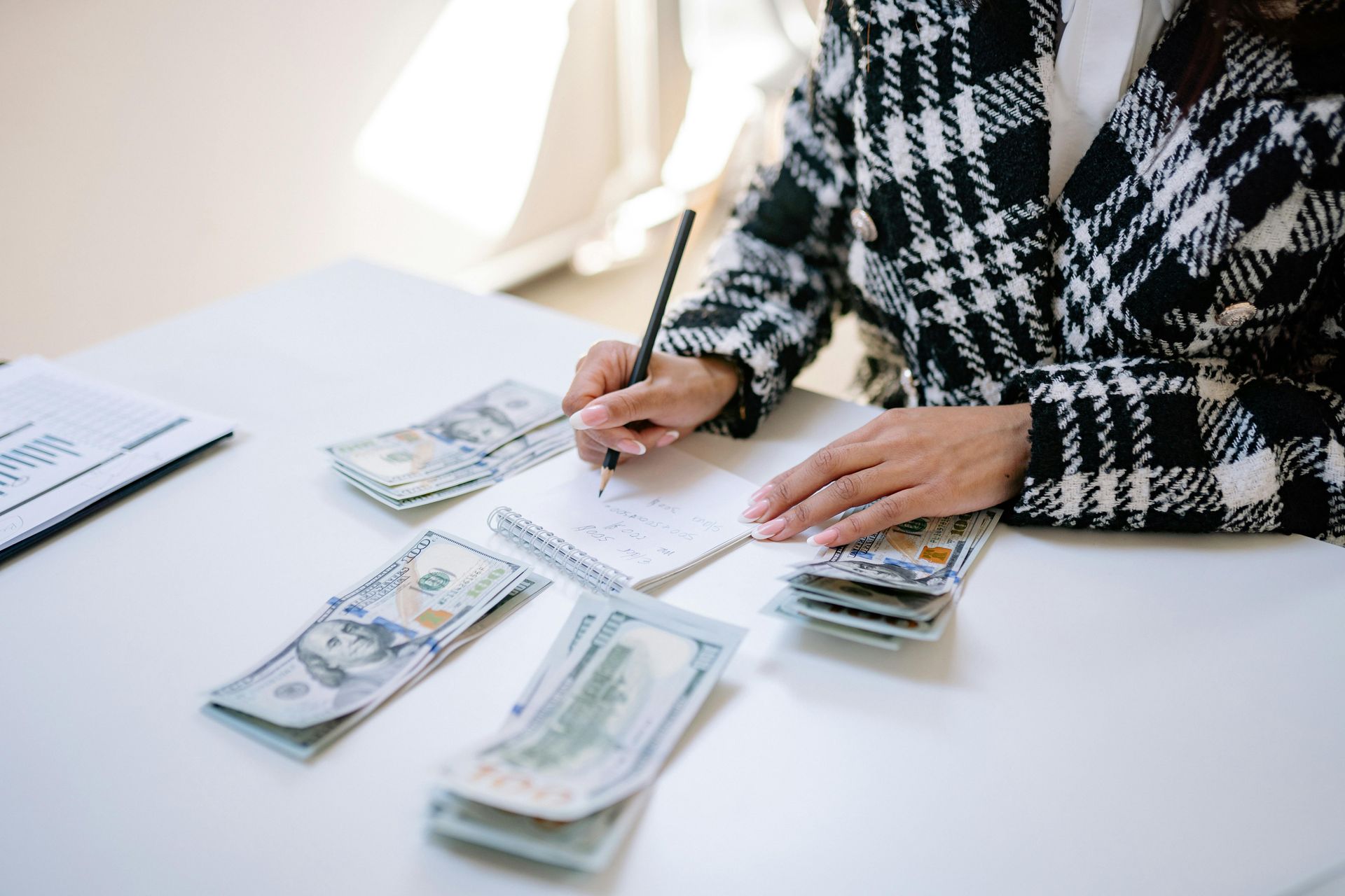 A person wearing a black and white plaid jacket sits at a white desk, writing on a notepad next to stacks of cash.