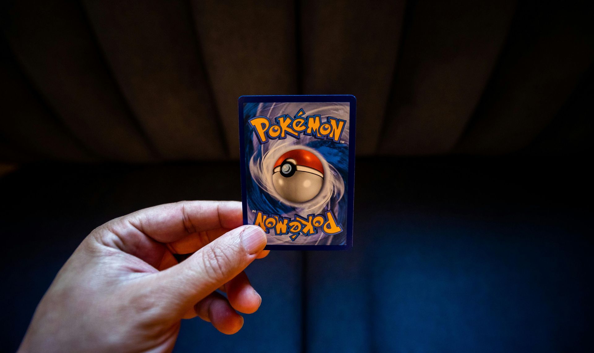 Hand holding a Pokémon trading card, displaying the blue back with a Pokéball. Dark background.