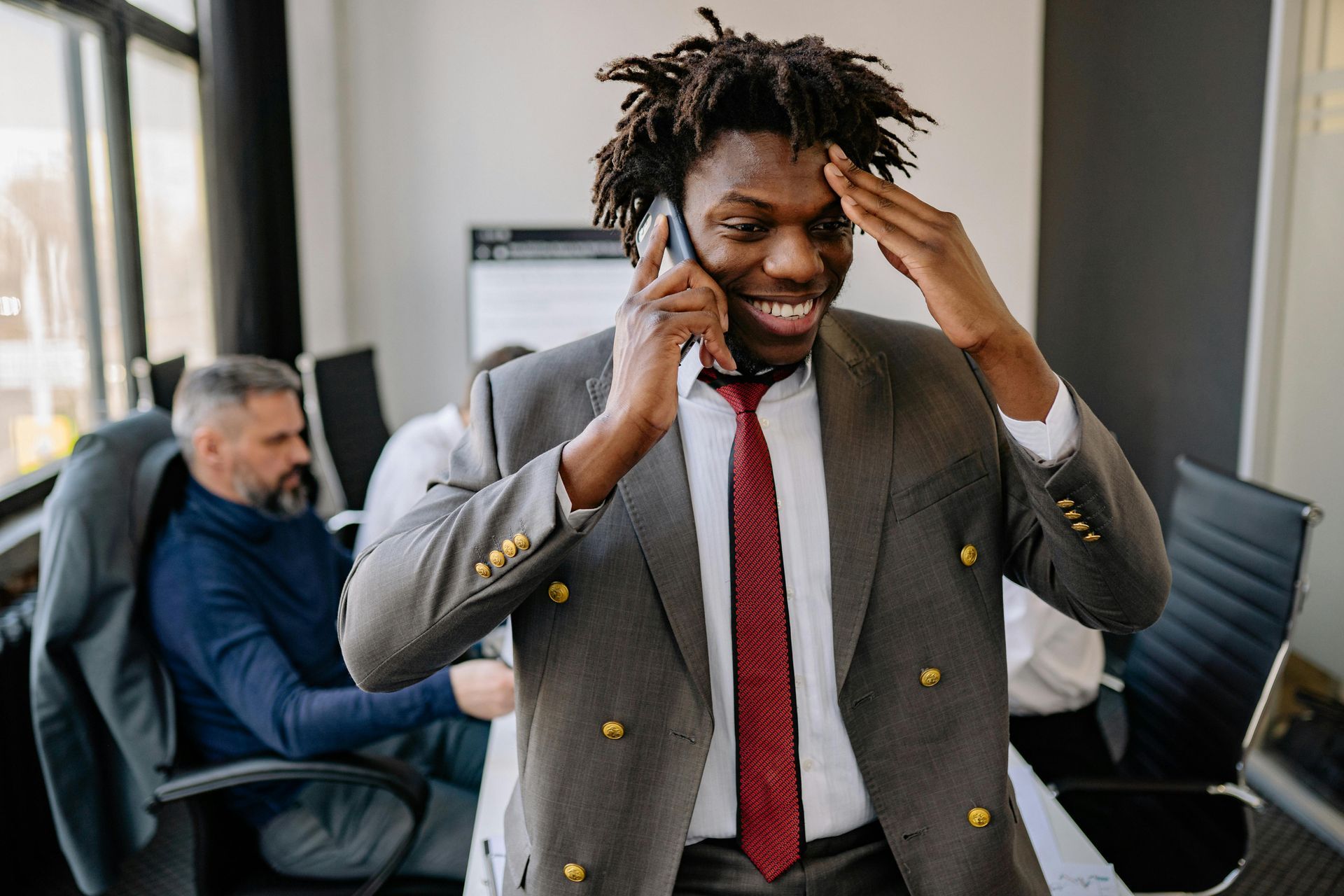 A smiling person in a suit and tie talks on a cell phone while holding a hand to their forehead in an office.