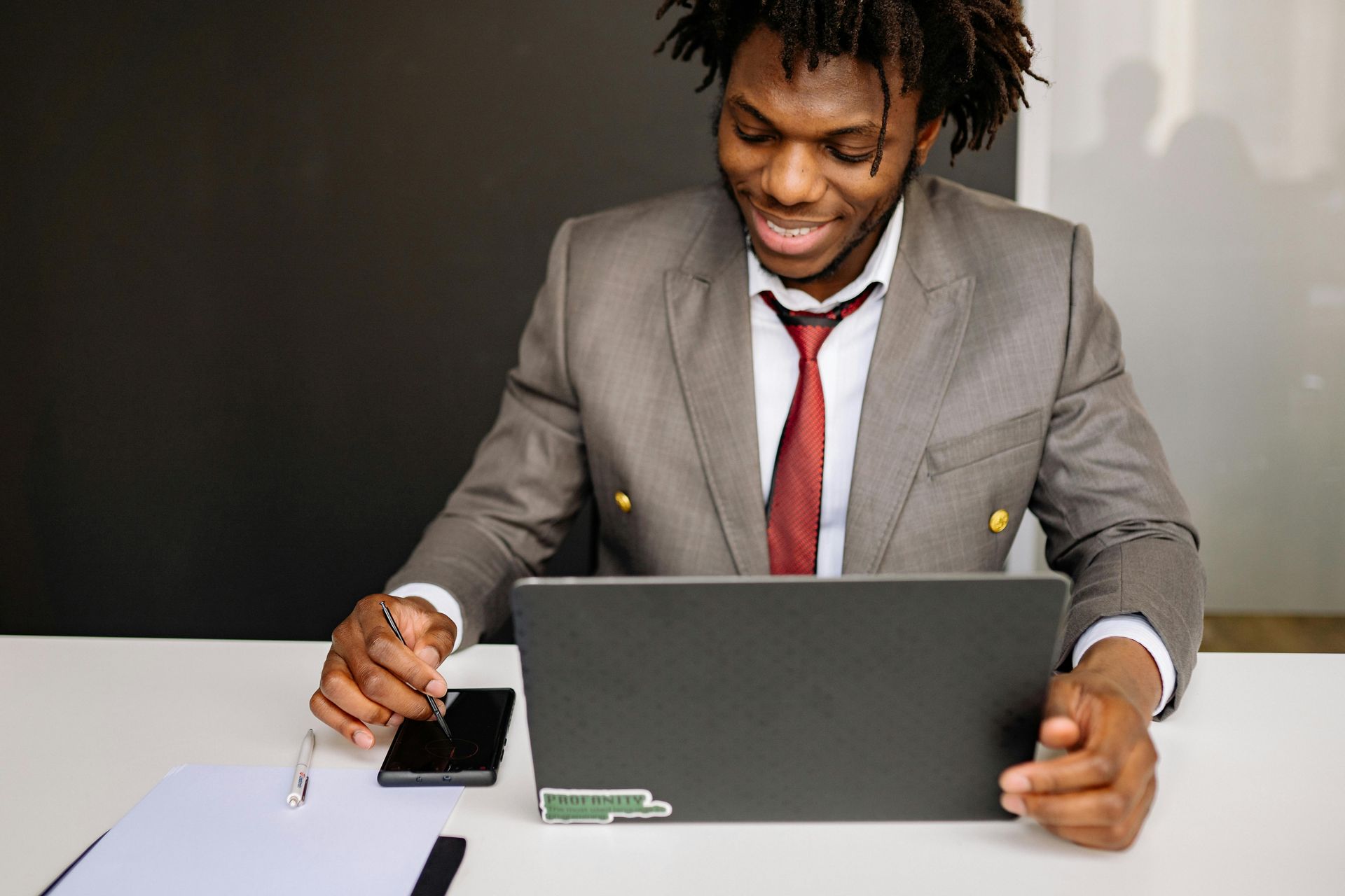 Man in gray suit at a desk, looking at a laptop, with phone and documents.
