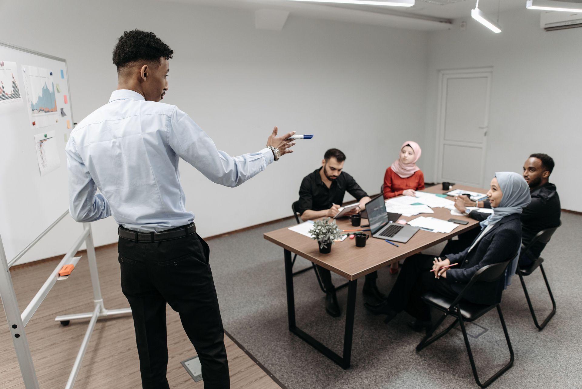 Presenter speaking to a small group in a meeting room with a whiteboard and laptop on the table.