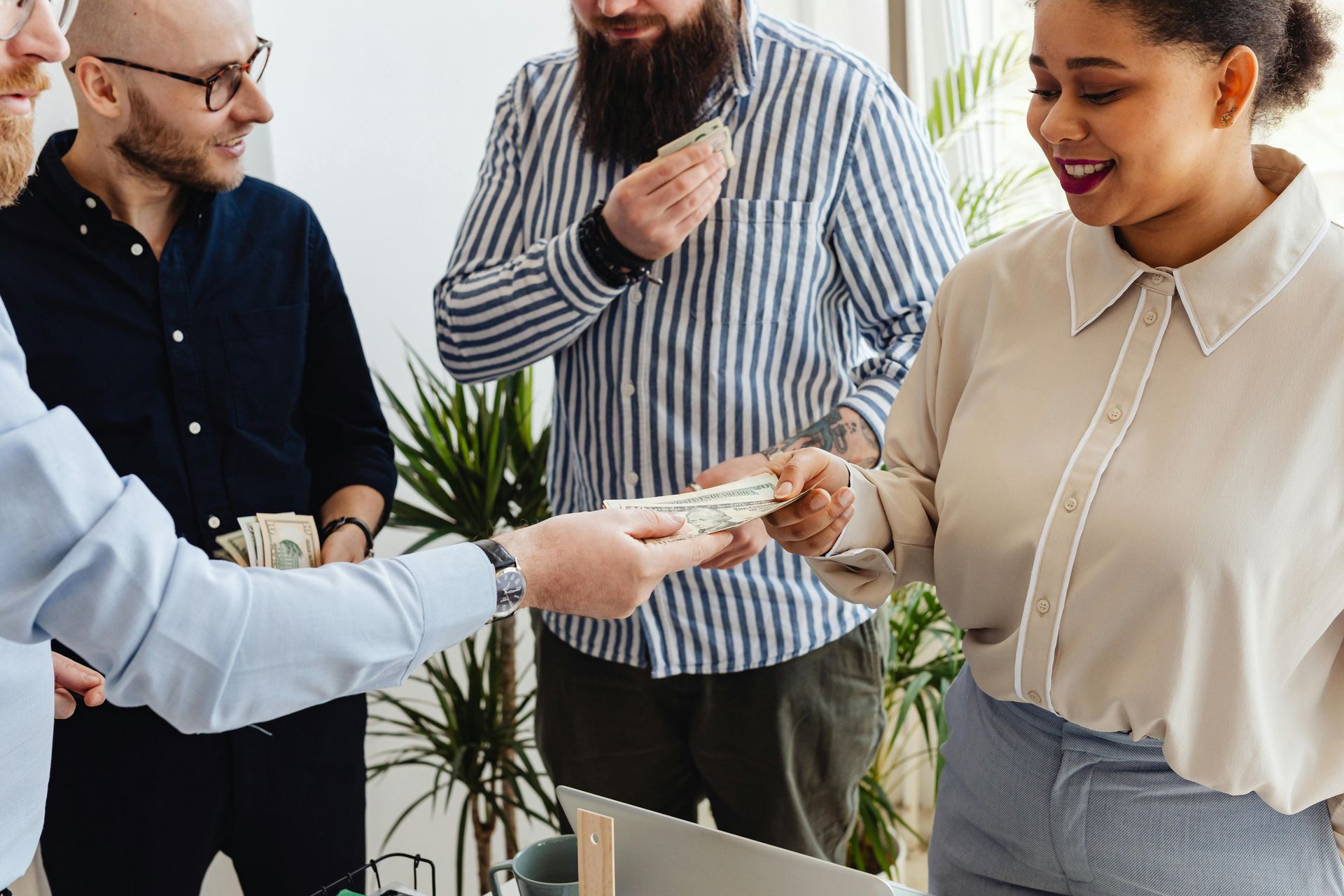 Four professionals gather around a table as one person hands a stack of cash to another in a bright office setting.
