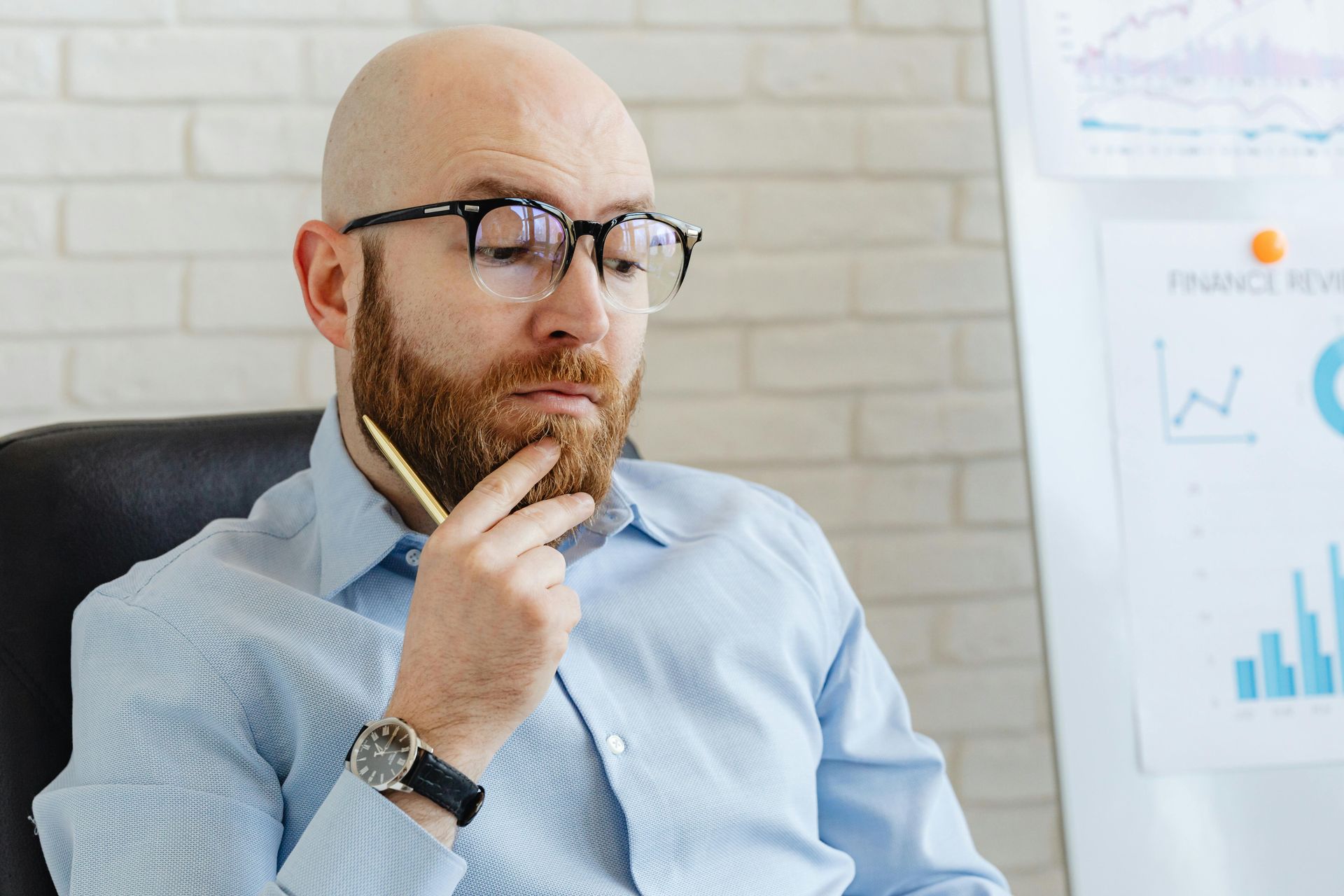 A man with a beard and glasses sits thoughtfully, holding a pen to his chin in an office with a financial chart.