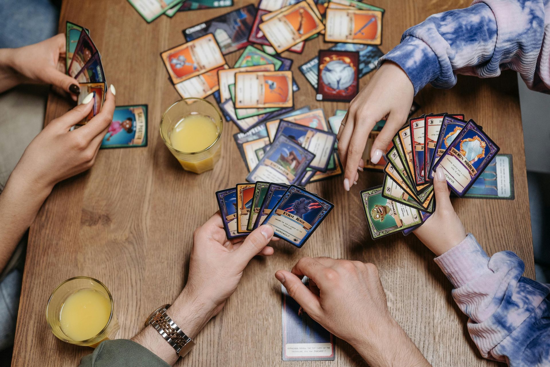 Three people play a tabletop card game on a wooden table with glasses of orange juice, holding cards in their hands.