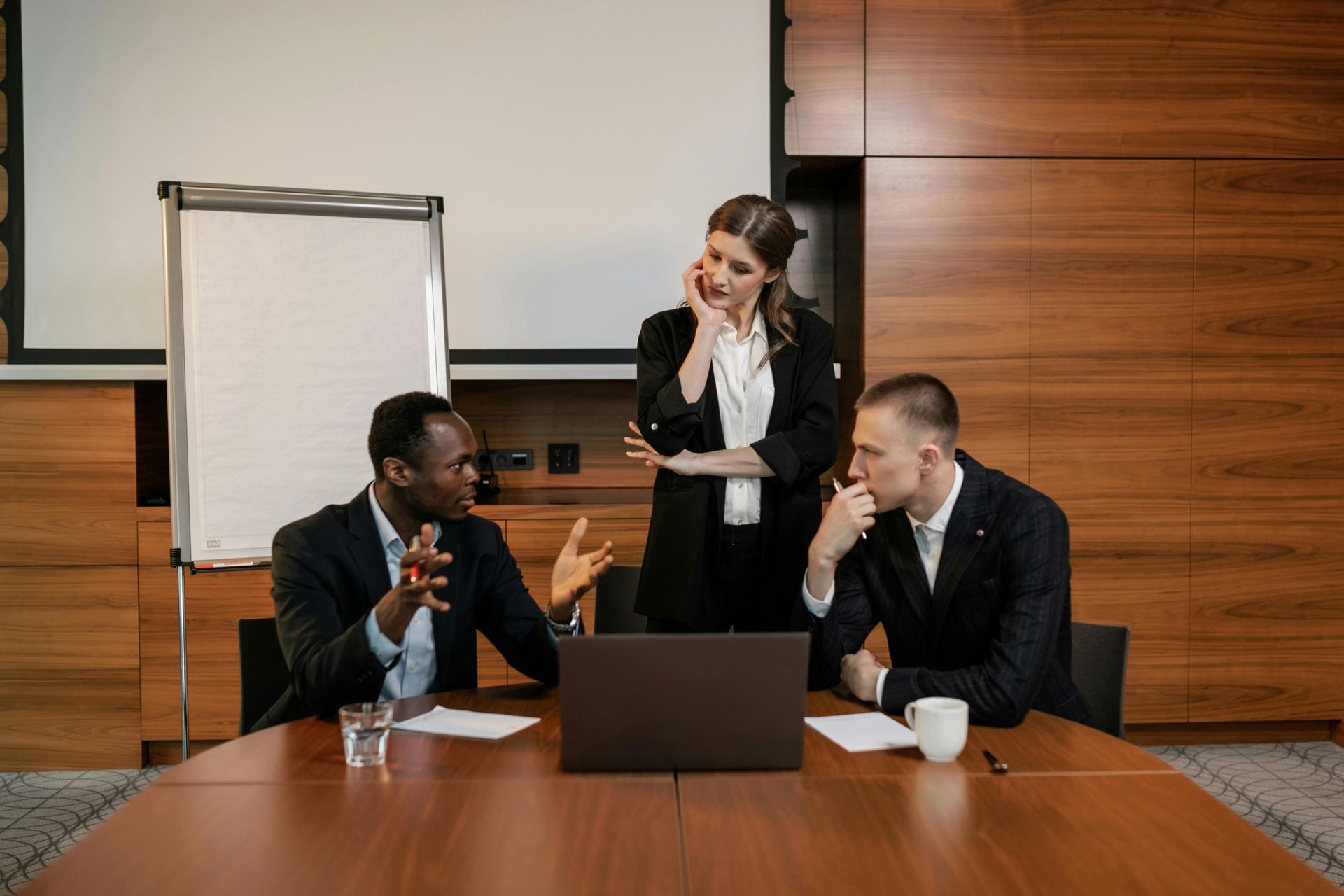 Three professionals in business attire collaborate around a laptop on a boardroom table.