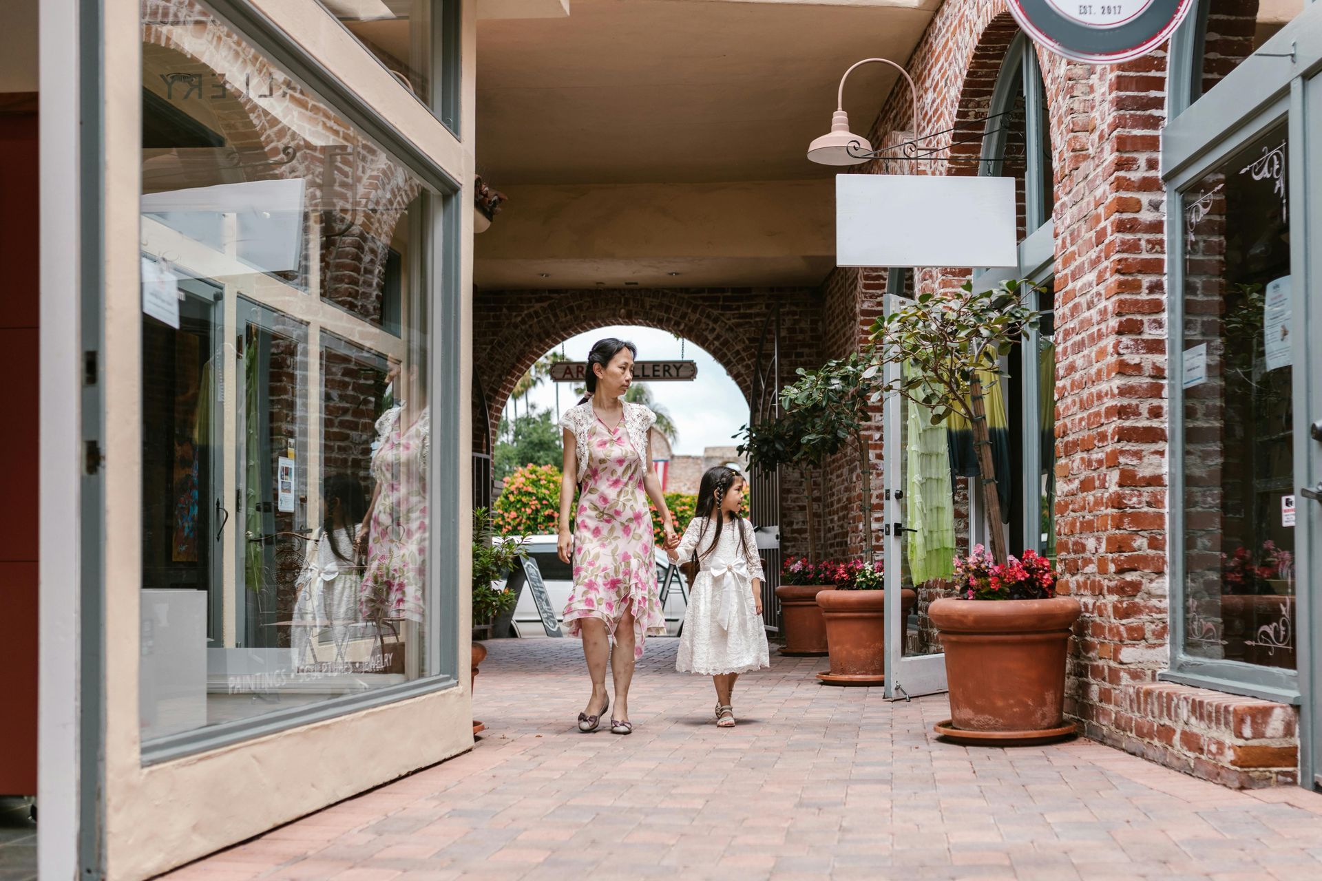 Woman and girl holding hands, walking in a brick-lined outdoor shopping area, past storefronts.