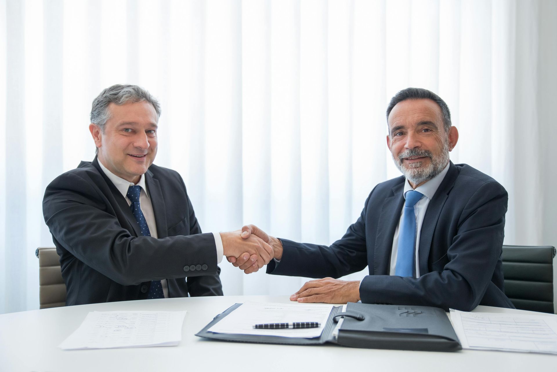 Two business professionals in suits shake hands over a desk with documents in a bright, modern office.