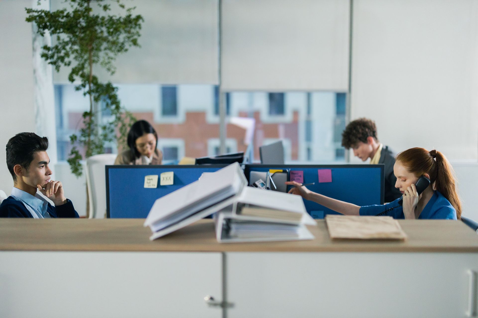 Office workers in a bright open-plan office, seated behind blue partitions with papers stacked in the foreground