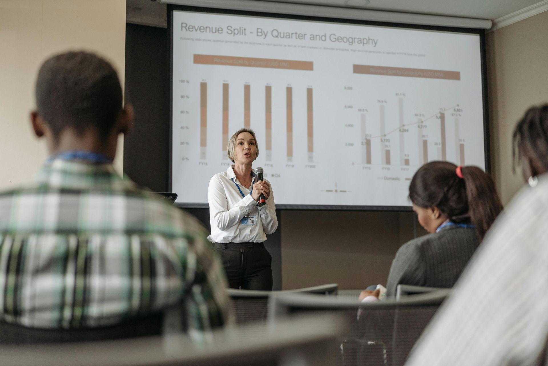 Woman presenting financial data in a meeting, using a microphone. Audience members seated, watching a slideshow.