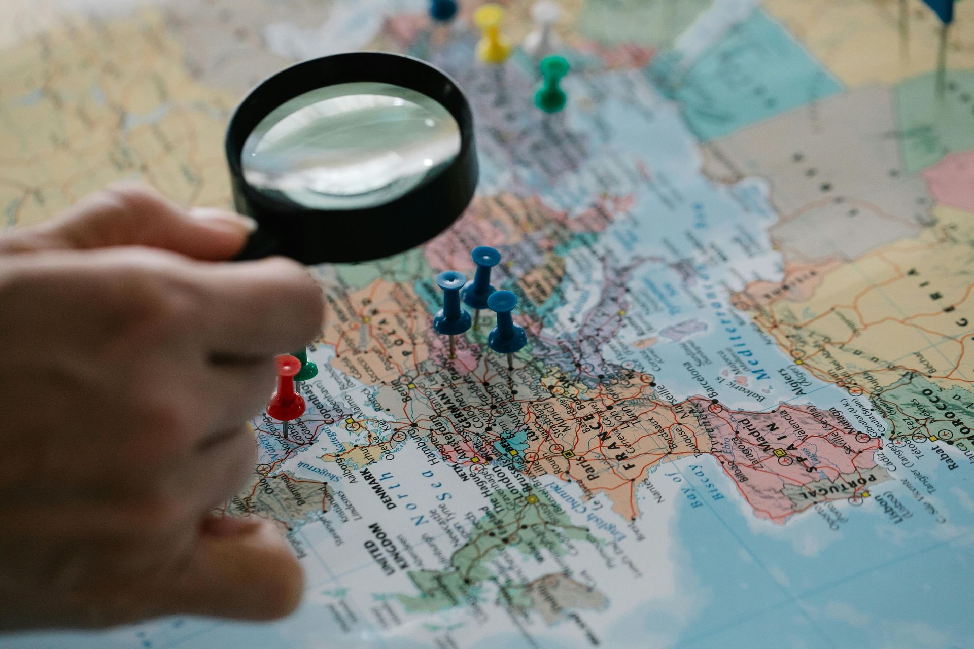 Hand holding a magnifying glass over a map with push pins marking locations.