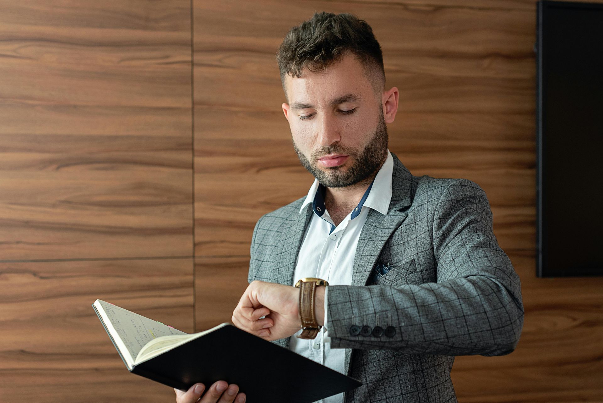 A person in a gray blazer and white shirt checks a wristwatch while holding an open notebook against a wood-paneled wall.