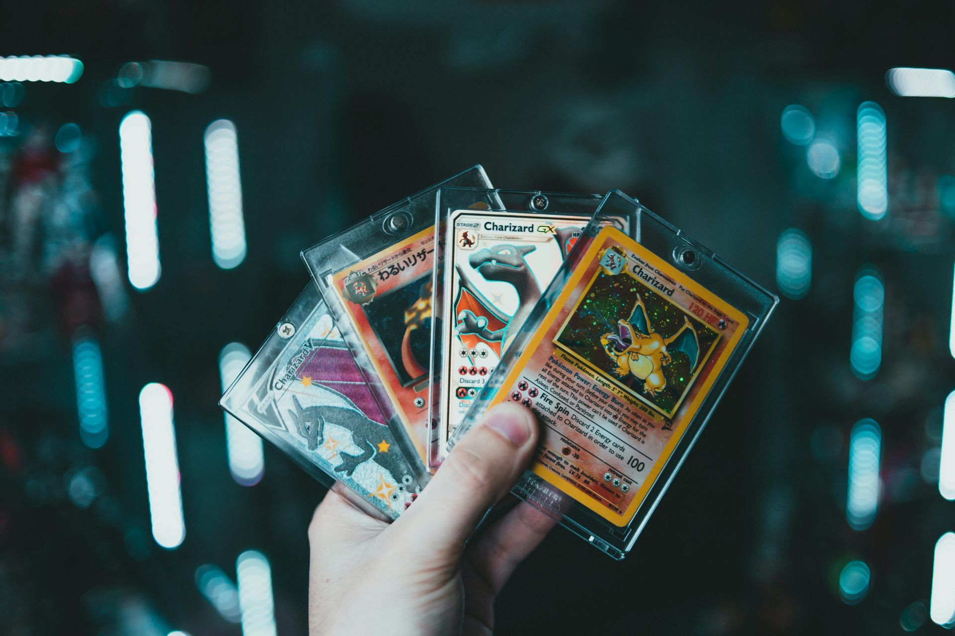 Hand holding four Pokémon cards in protective sleeves, in front of a blurred background of lights.
