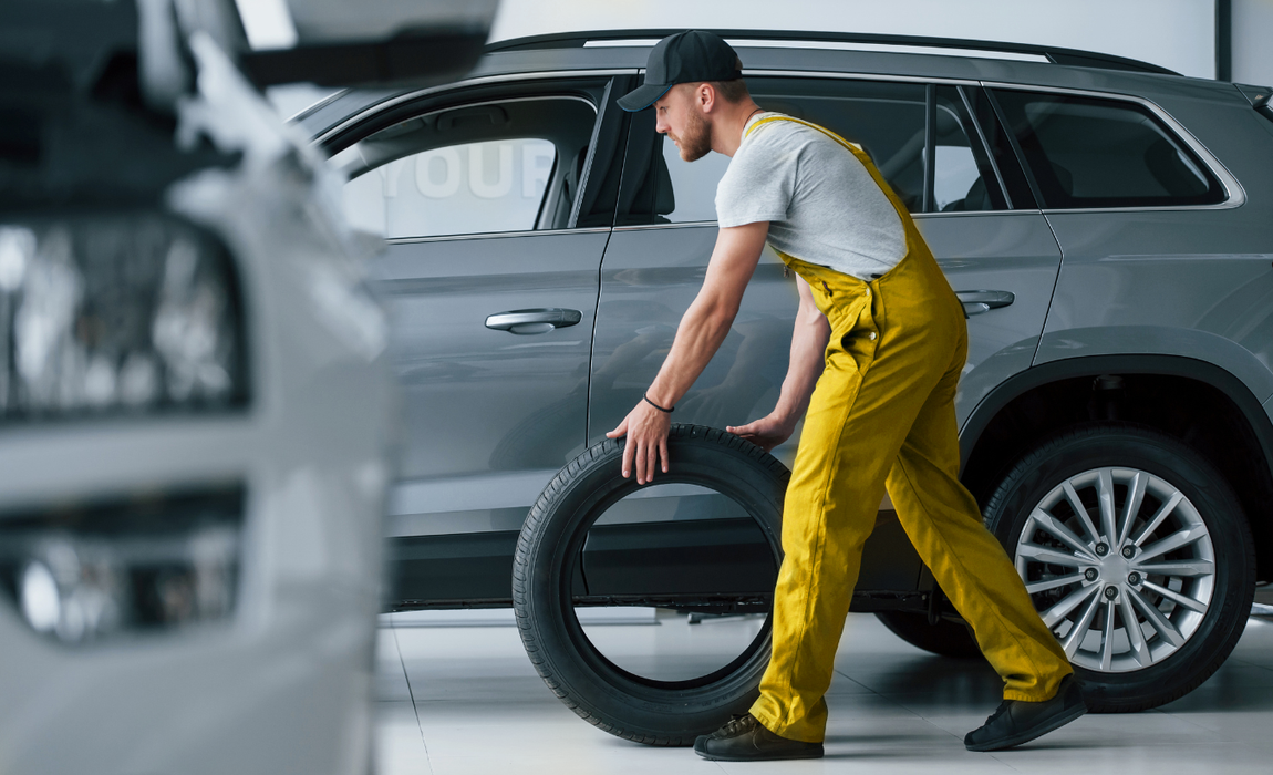 Mecânico de macacão amarelo empurrando um pneu em direção a um SUV cinza em uma oficina.