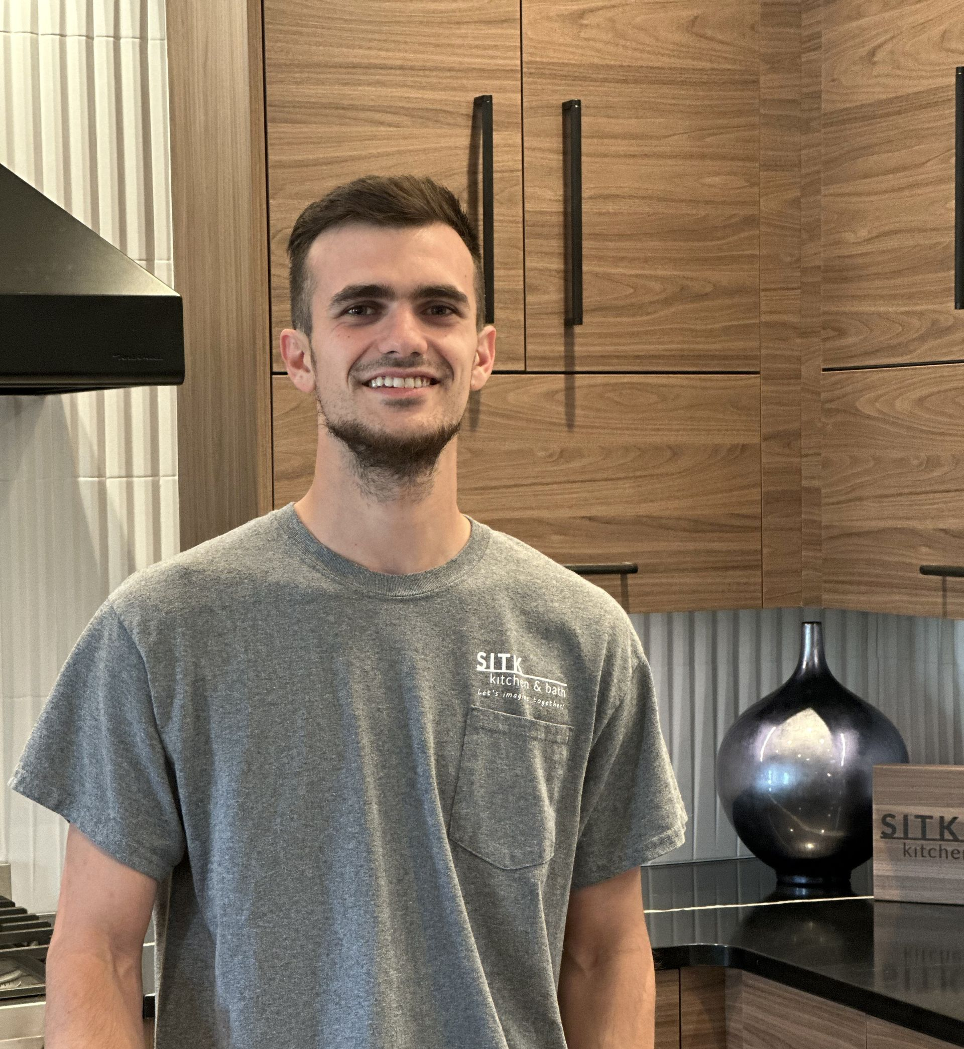 Smiling man in a gray T-shirt standing in a modern kitchen with wood cabinets.