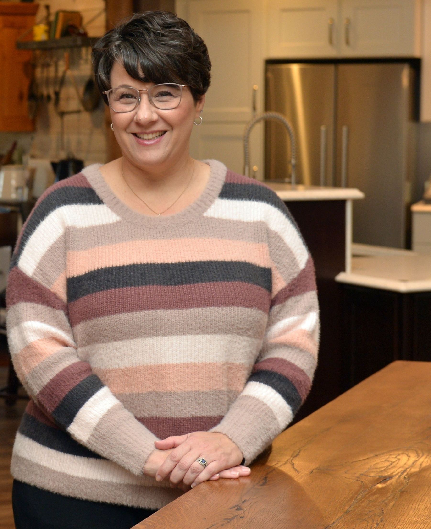 Smiling person in a striped sweater standing in a kitchen with hands clasped.