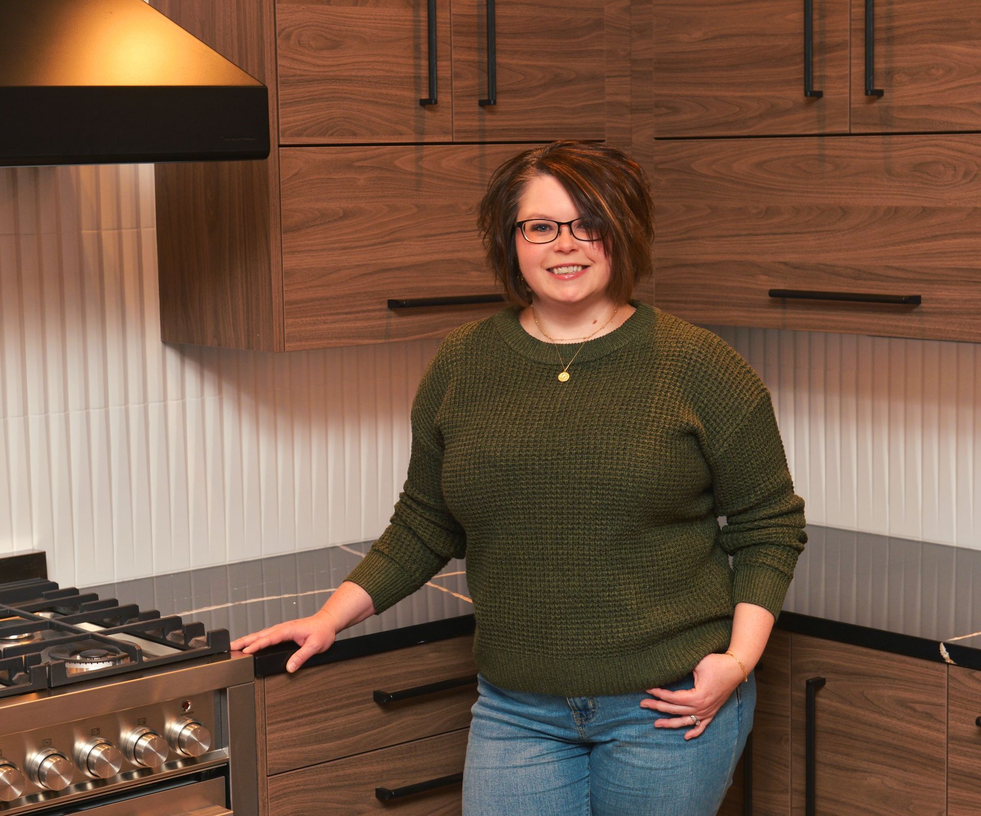 Person standing in a modern kitchen, smiling beside a stove and wood cabinets.