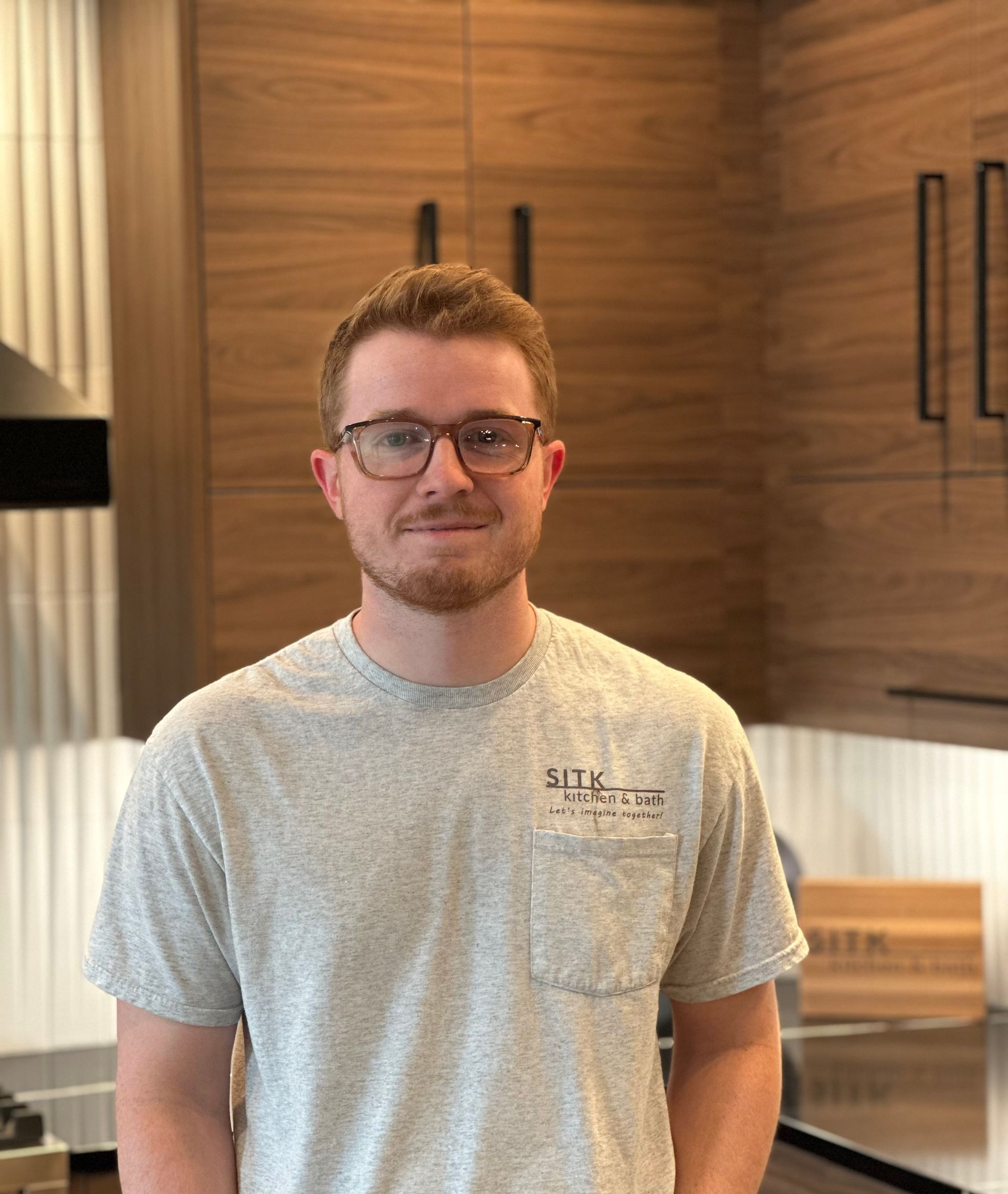 Person standing indoors in a kitchen, wearing glasses and a light gray T-shirt, with wood cabinets behind them.