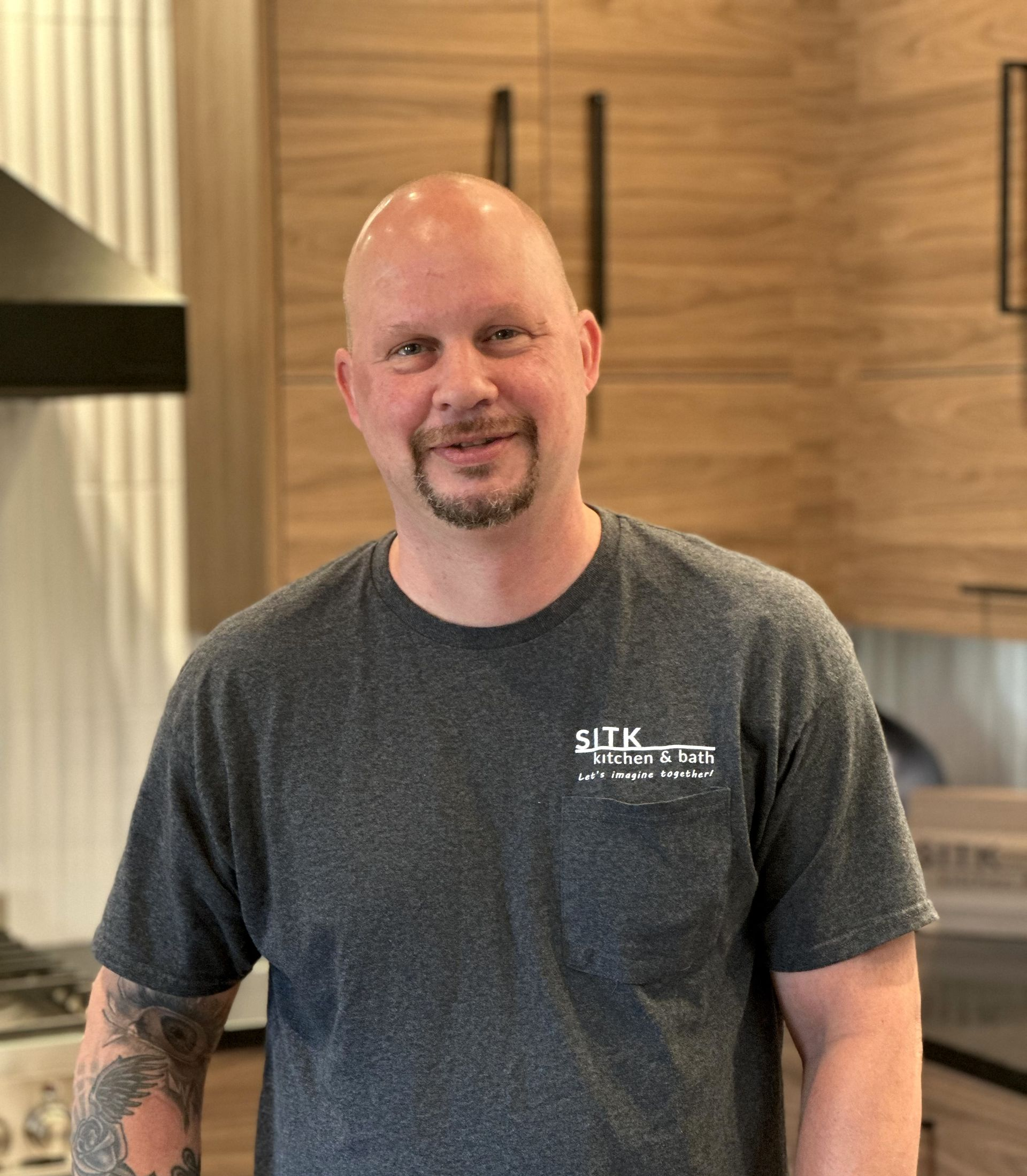 Man standing indoors in a kitchen, wearing a gray T-shirt, with wood cabinets in the background.