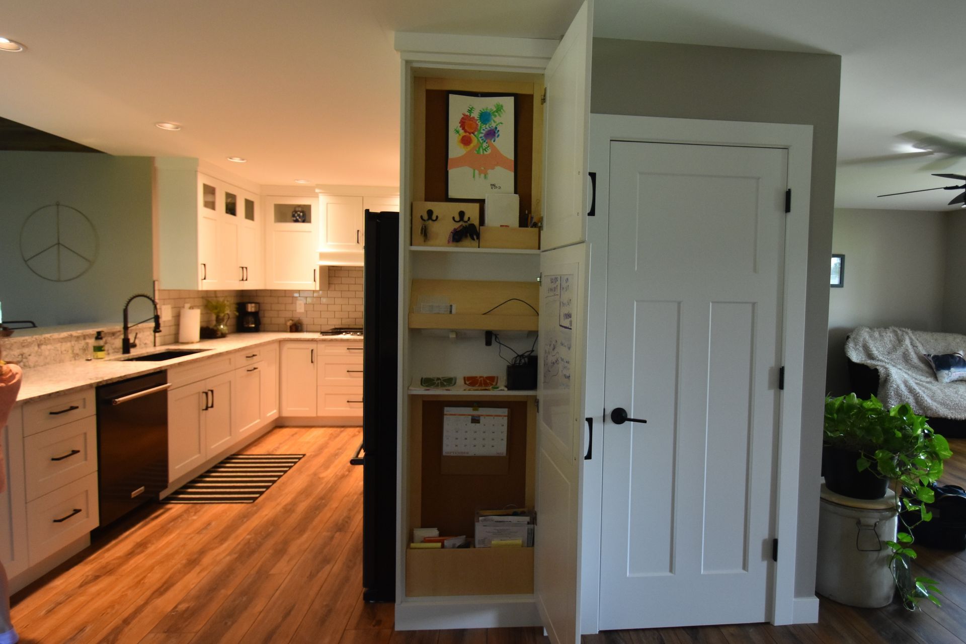 Open kitchen and living area with white cabinets, wood floor, and a narrow shelving unit by a doorway.