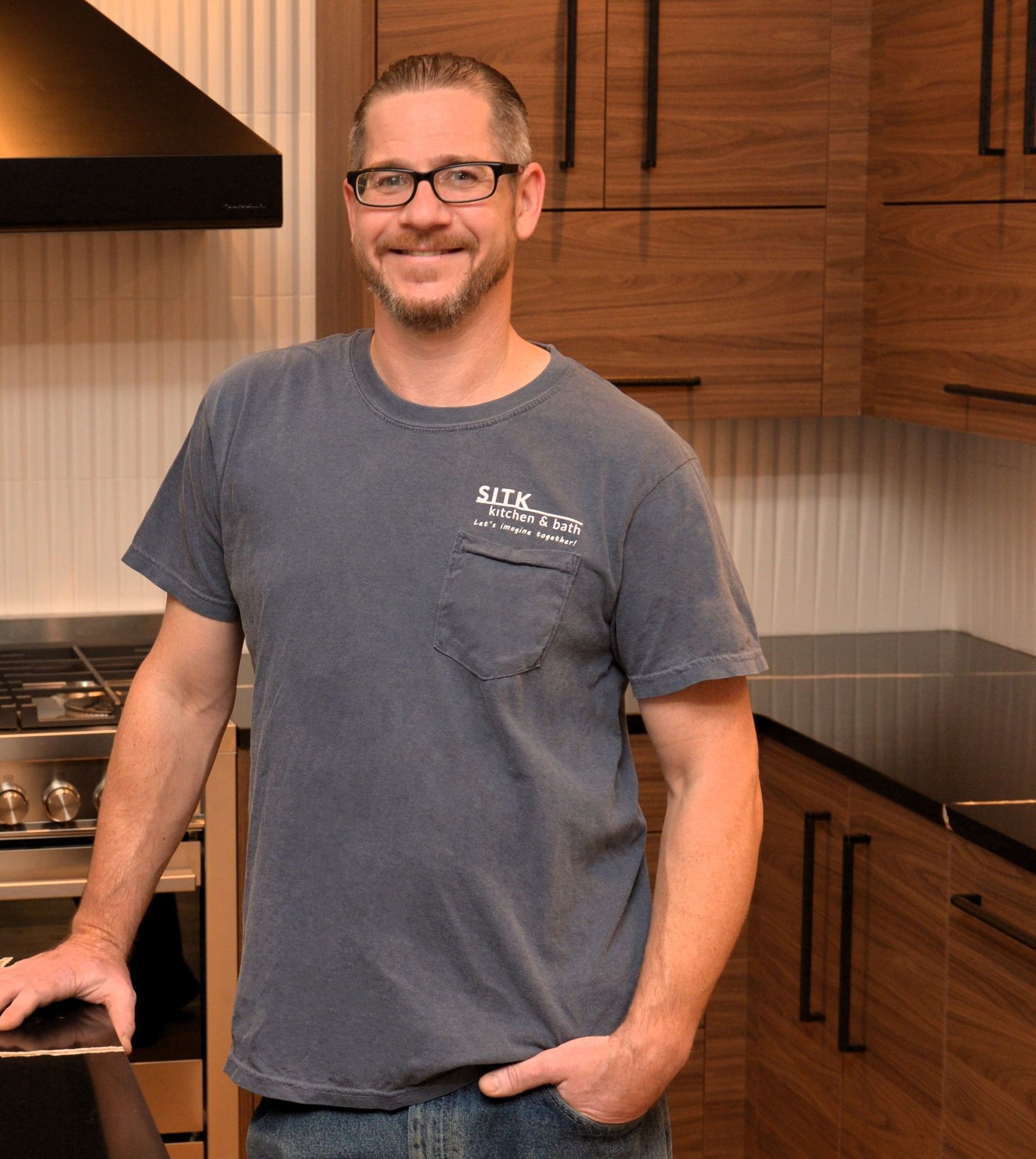 Man standing in a modern kitchen, leaning on the counter and smiling, wearing a gray T-shirt.