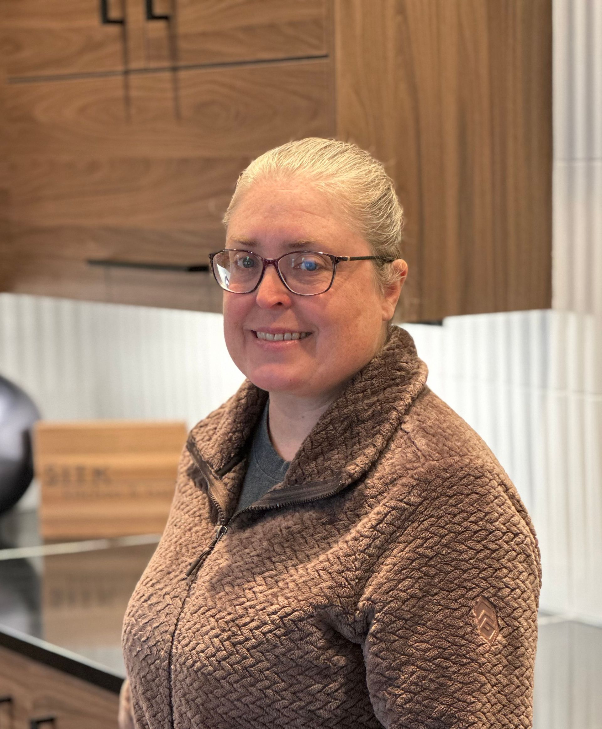Person smiling in a brown fleece sweater, standing in a modern kitchen with wood cabinets and white tile.