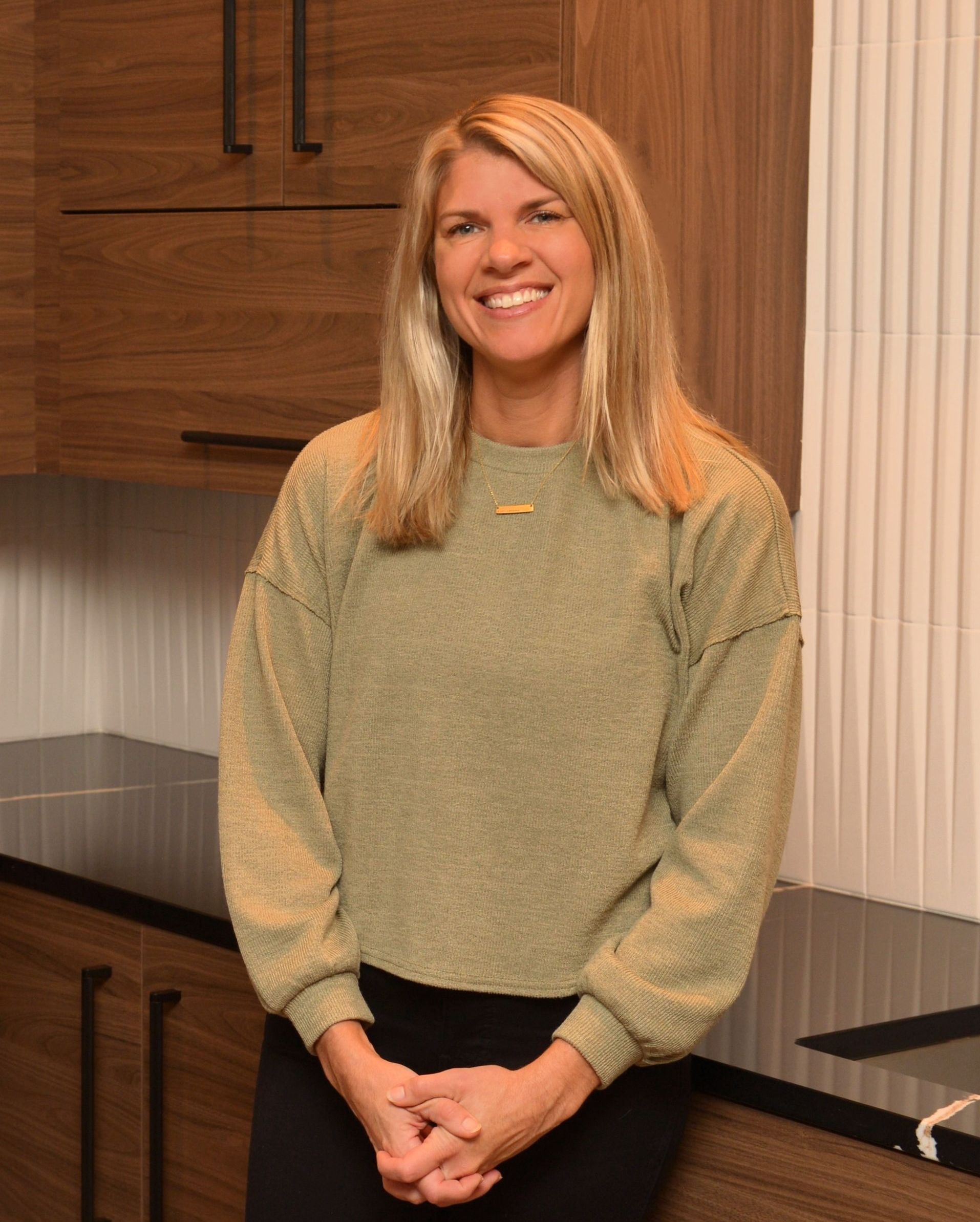 Smiling woman in a kitchen, wearing a light green sweater and standing by dark wood cabinets.