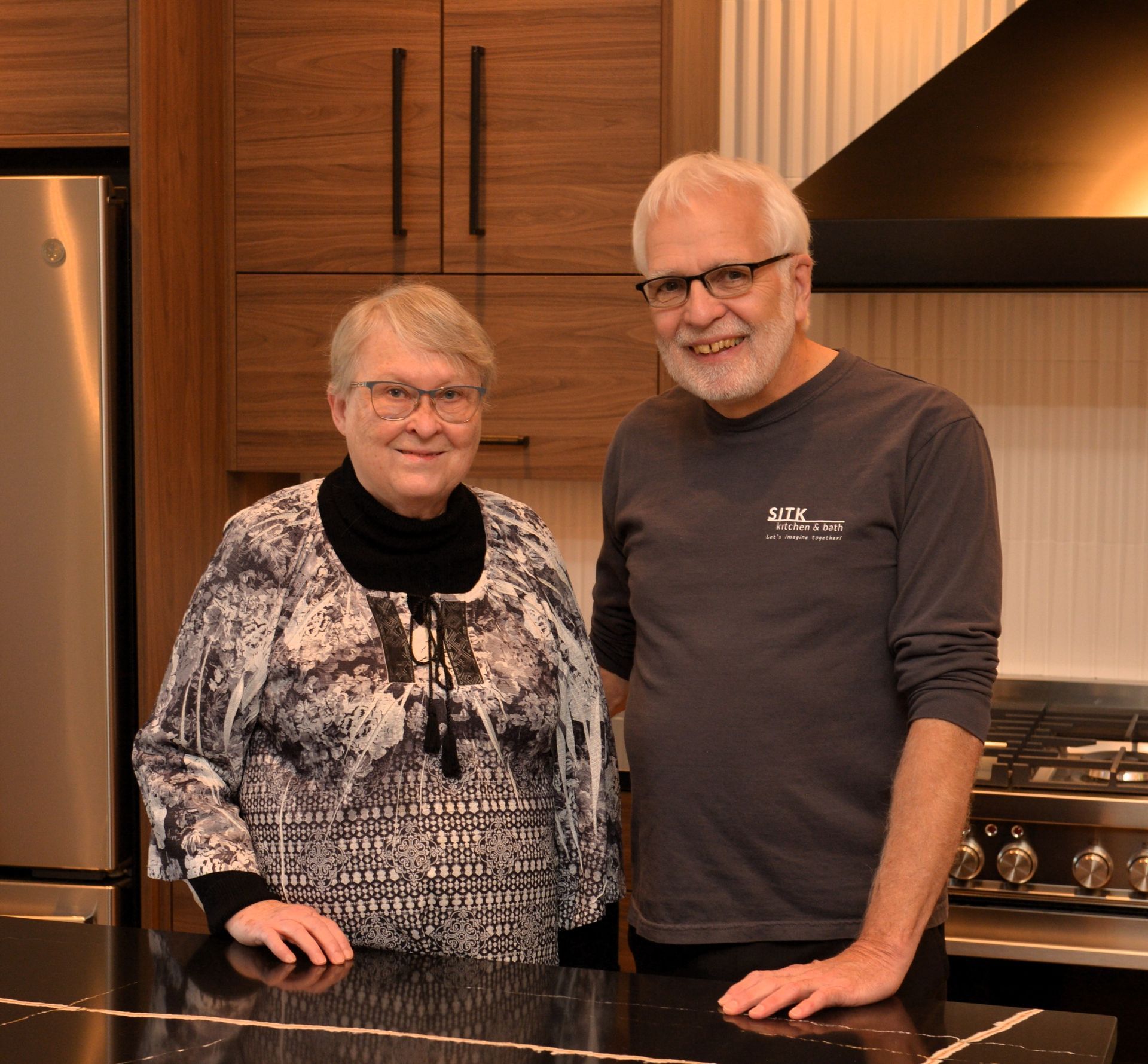 Two people smiling in a modern kitchen, standing behind a black countertop.