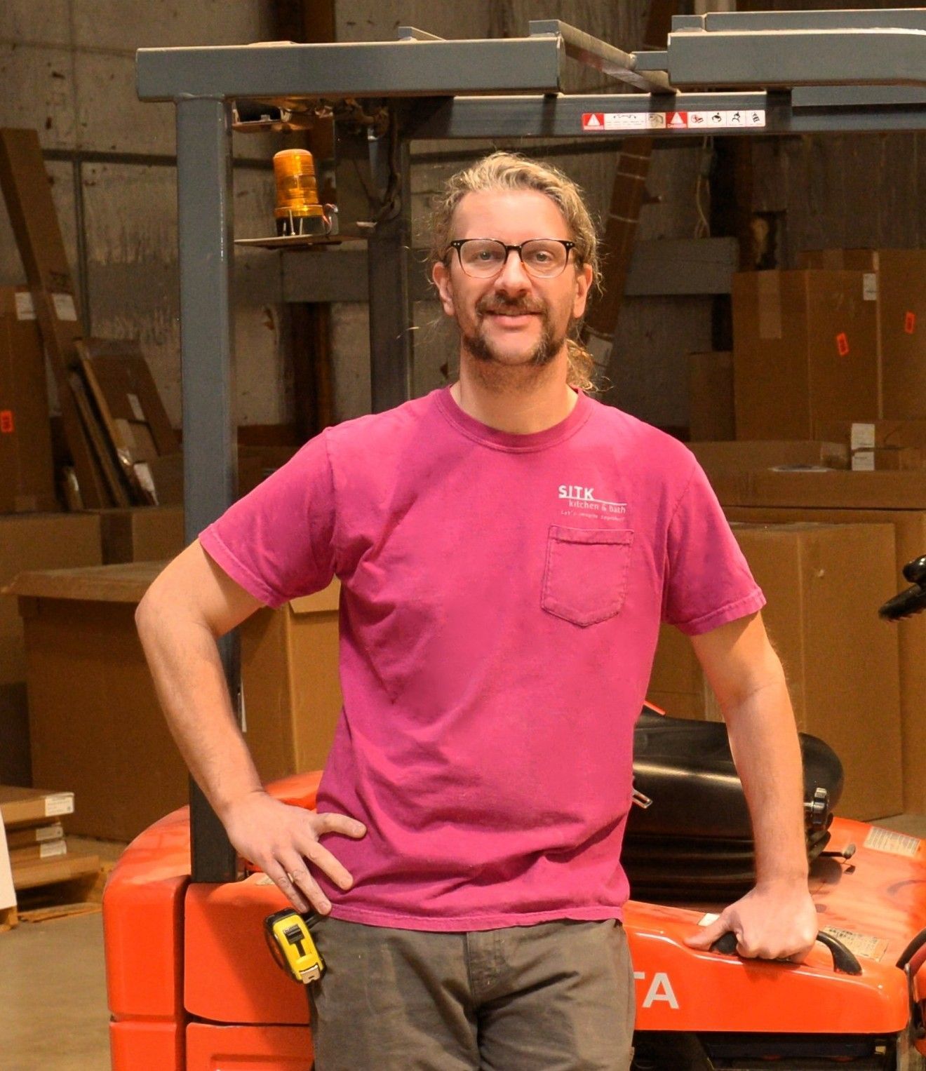 Man standing with hands on hips beside an orange forklift in an industrial warehouse.