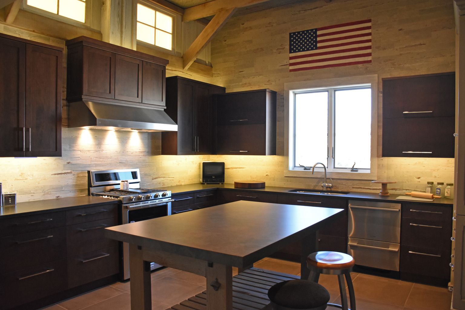 Modern kitchen with dark cabinets, central island, wood beams, window, and American flag on the wall