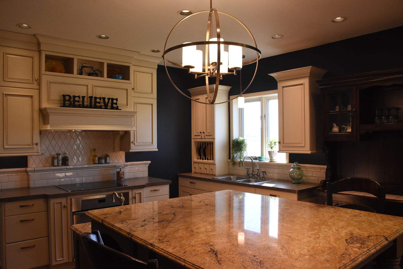 Spacious kitchen with granite island, cream cabinets, and a hanging chandelier by a bright window.